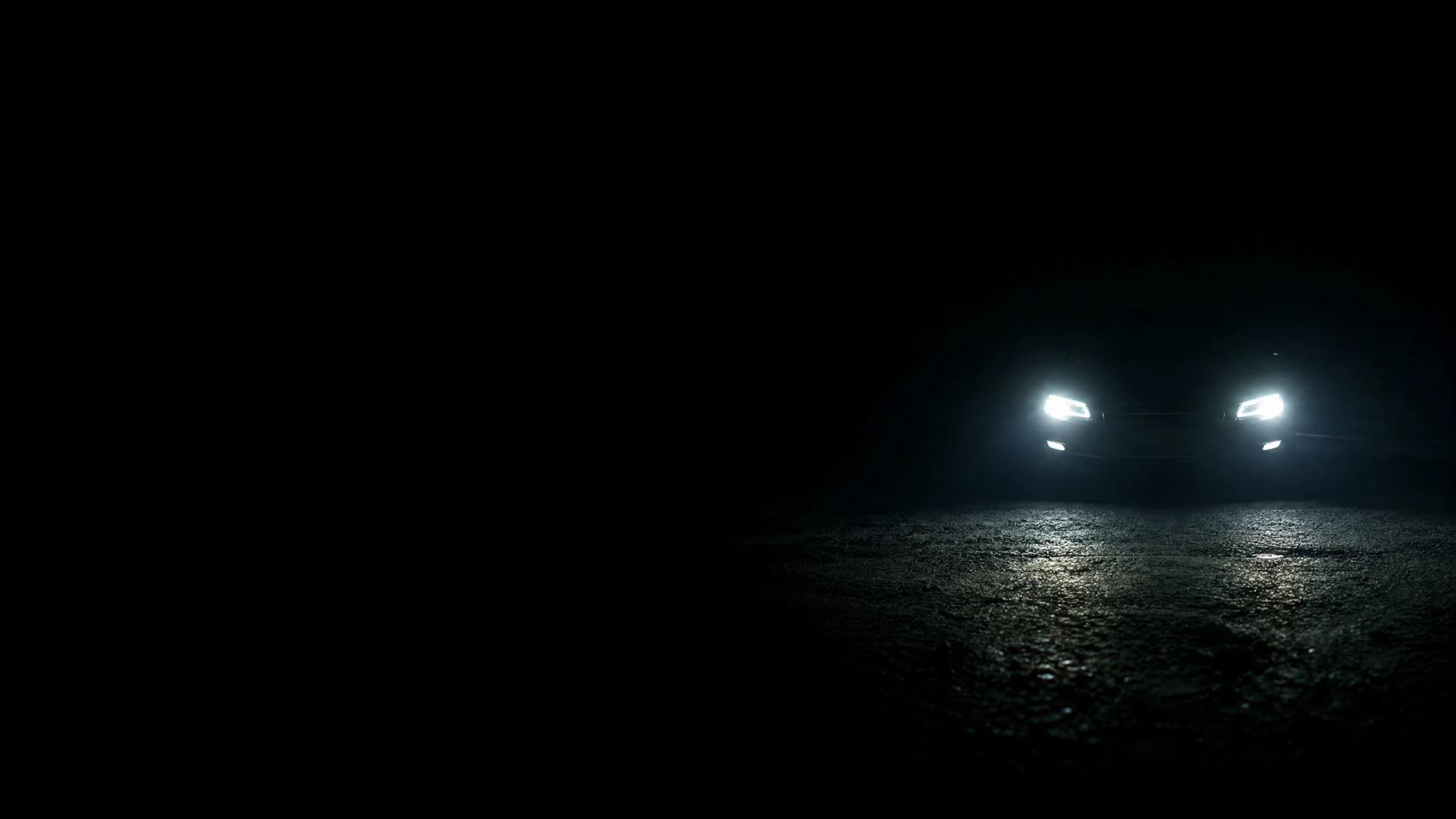 A car sits on a dark road at night with two bright headlights casting light onto the ground below.