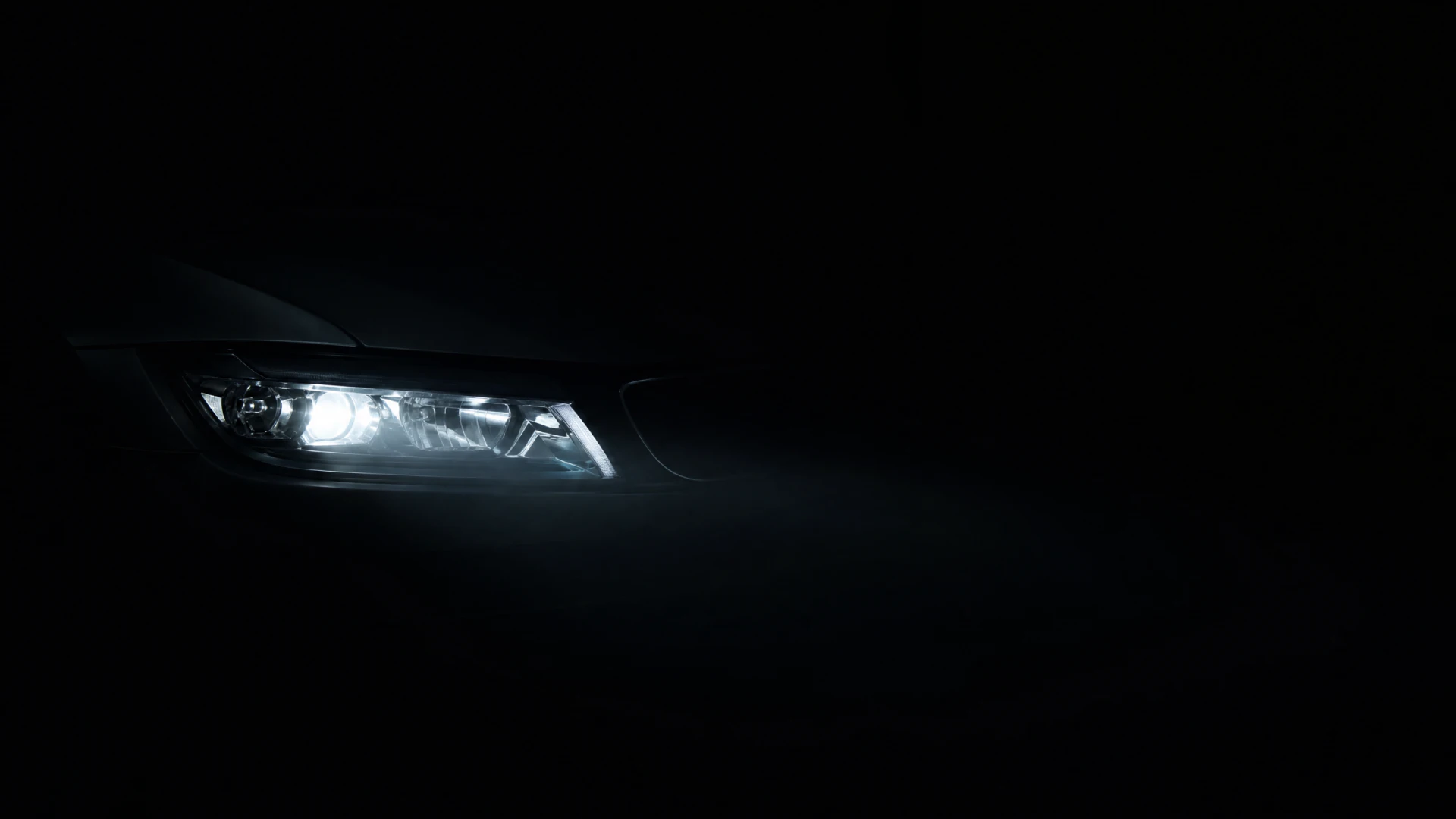 A single illuminated car headlight with a projector lens and angular LED accent lights, photographed against a black background.