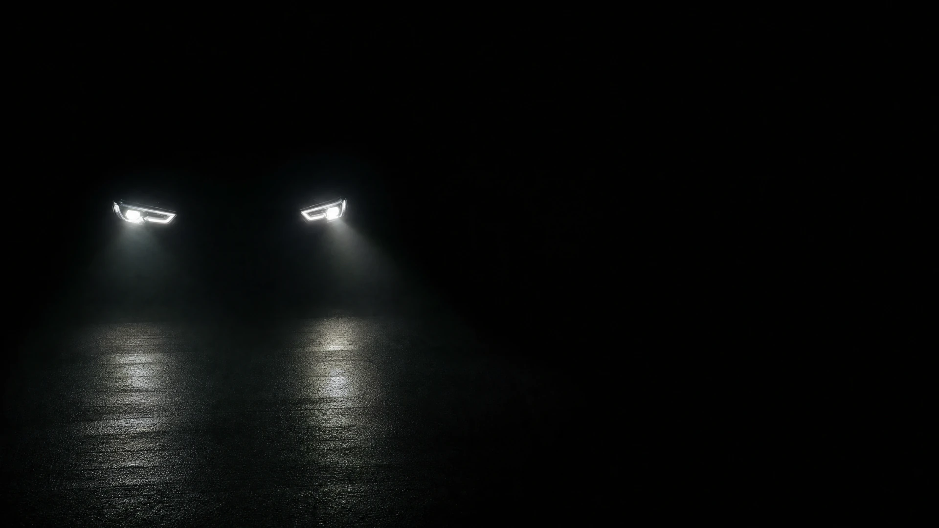 Two glowing LED headlights shine downward onto a wet dark road surface, photographed from the front in near-total darkness.
