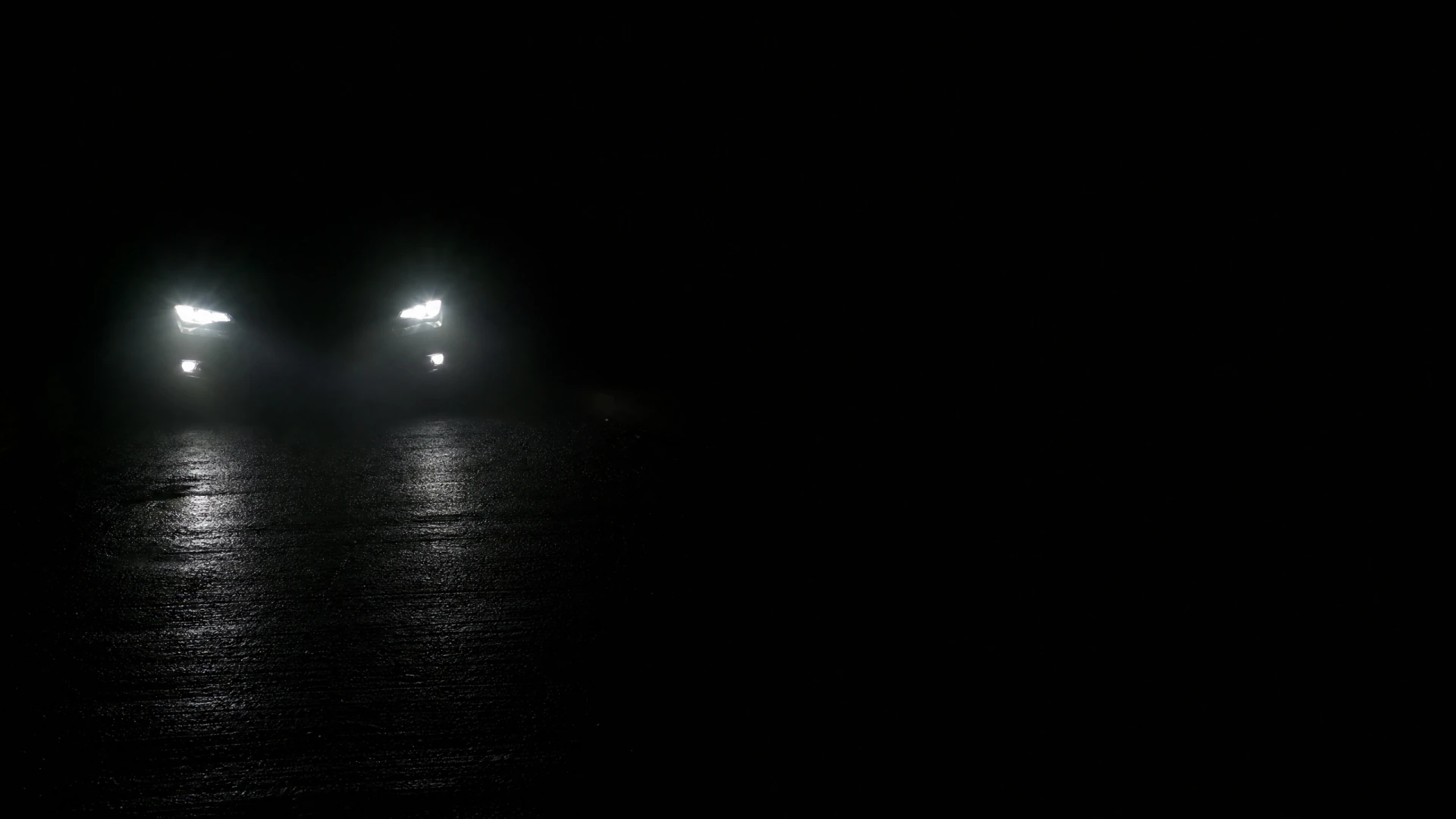 A vehicle faces the camera at night, its two bright white headlights casting reflections on a wet road surface.