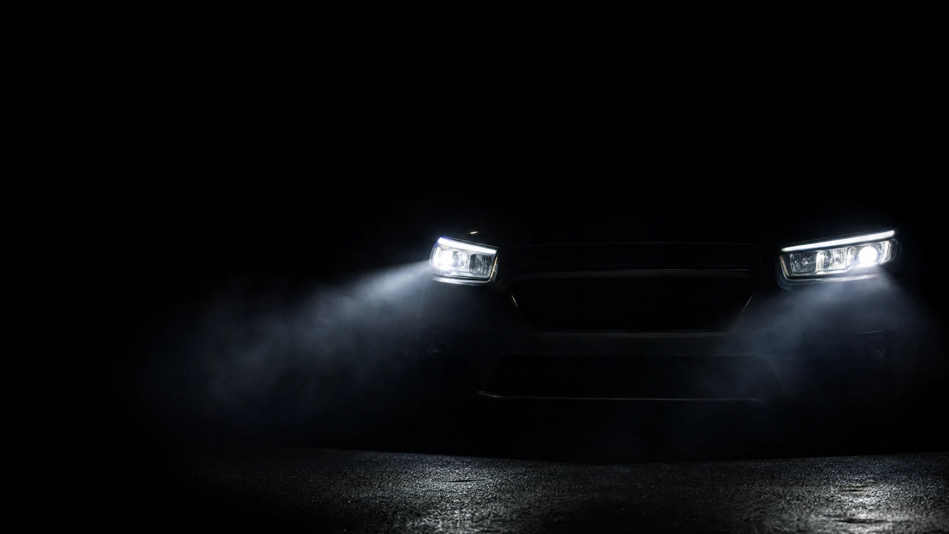 The illuminated headlights of a dark vehicle shine through low-lying mist on a wet surface at night.