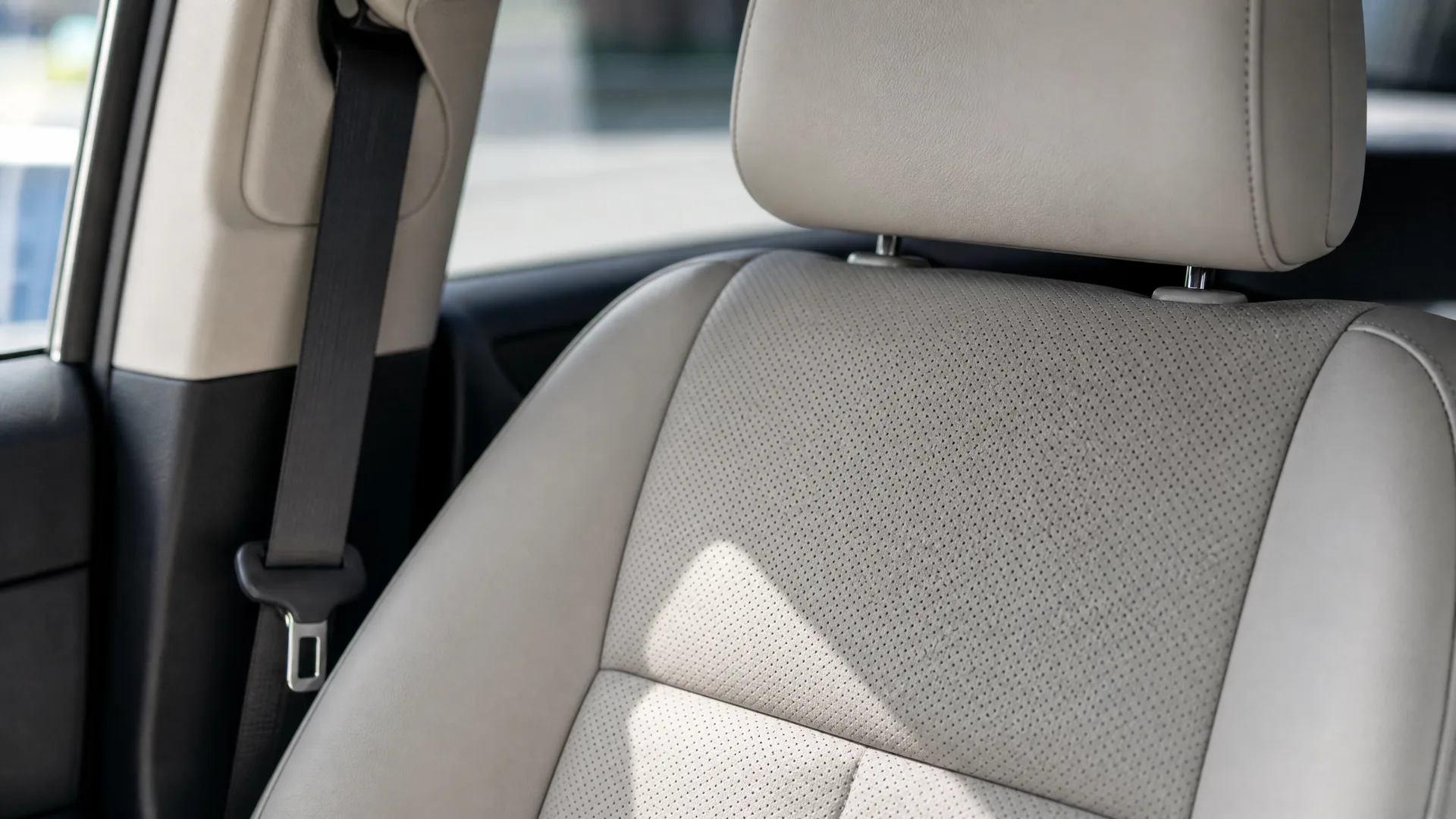 Close-up of a beige perforated leather car front seat with headrest, a black seatbelt and window visible at left.