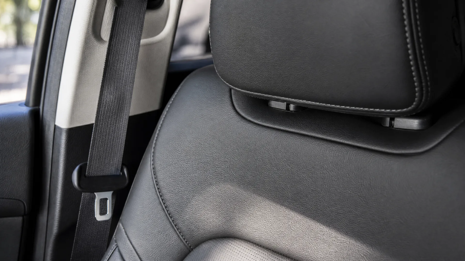 Close-up of a black leather car seat back and headrest with a seat belt and door pillar visible at left, blurred greenery through the window.