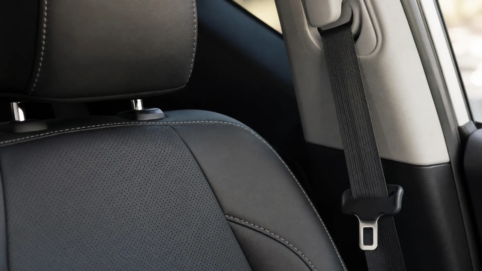 Close-up of a black leather car seat with headrest beside a B-pillar with a black seat belt and metal buckle tongue.
