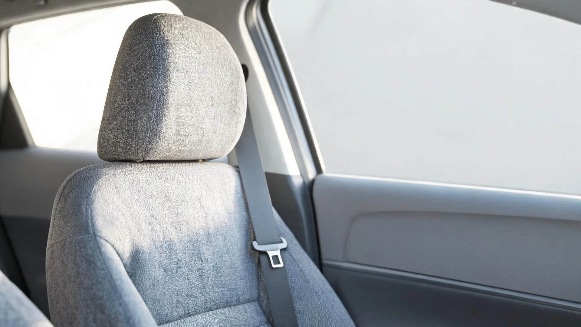 Close-up of a gray fabric car front seat with headrest and seat belt beside a door panel and window inside a vehicle.