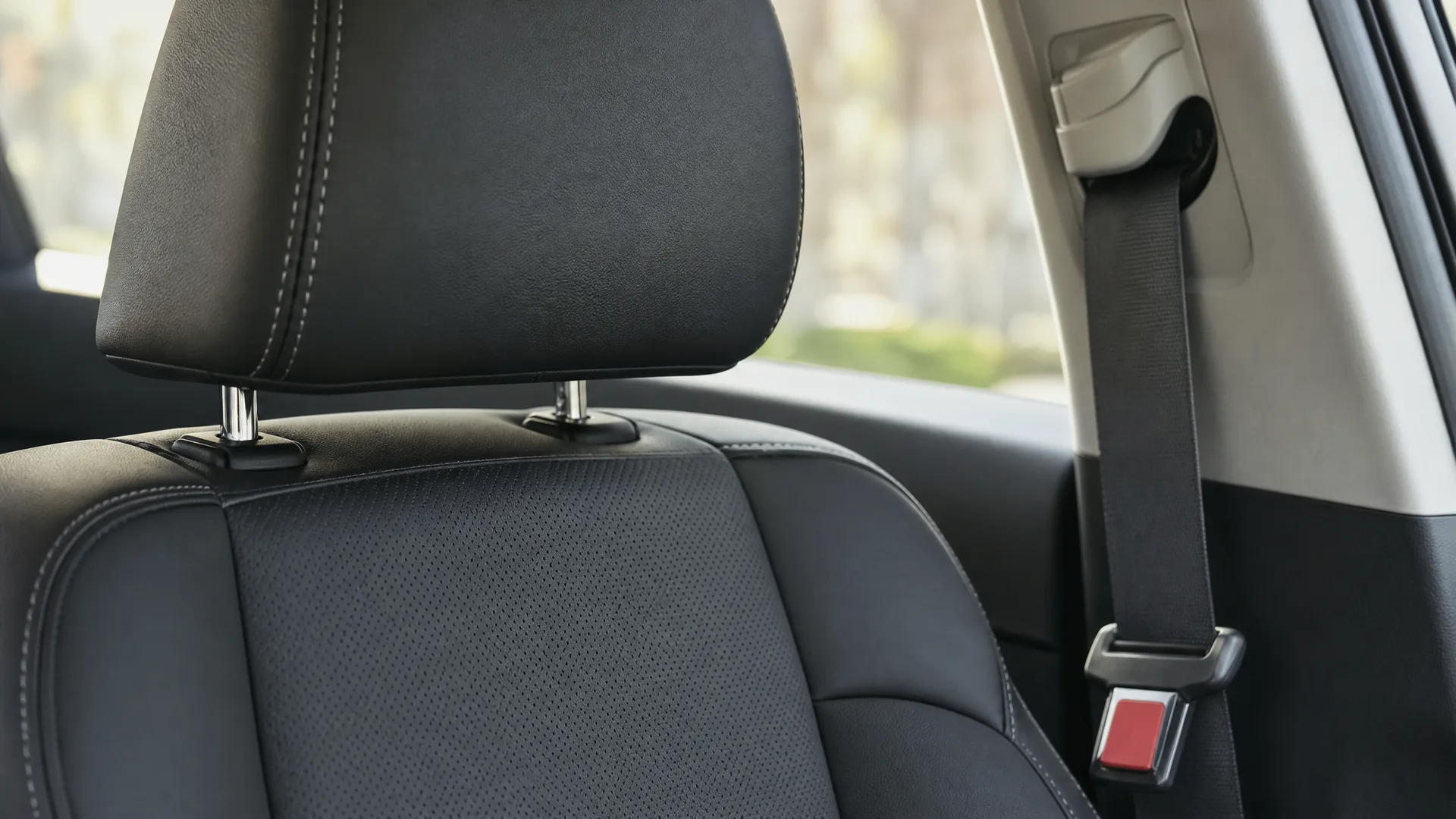 Close-up of a black leather car seat with headrest beside a window and a seat belt with red buckle release, with blurred greenery outside.