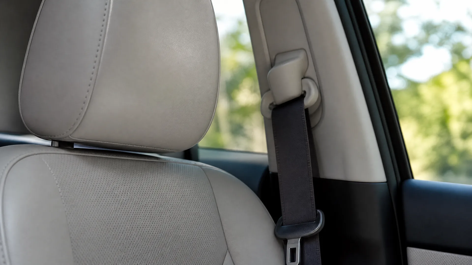 Close-up of a beige leather car seat and headrest beside a dark seat belt on the B-pillar, with blurred green trees visible through the window.