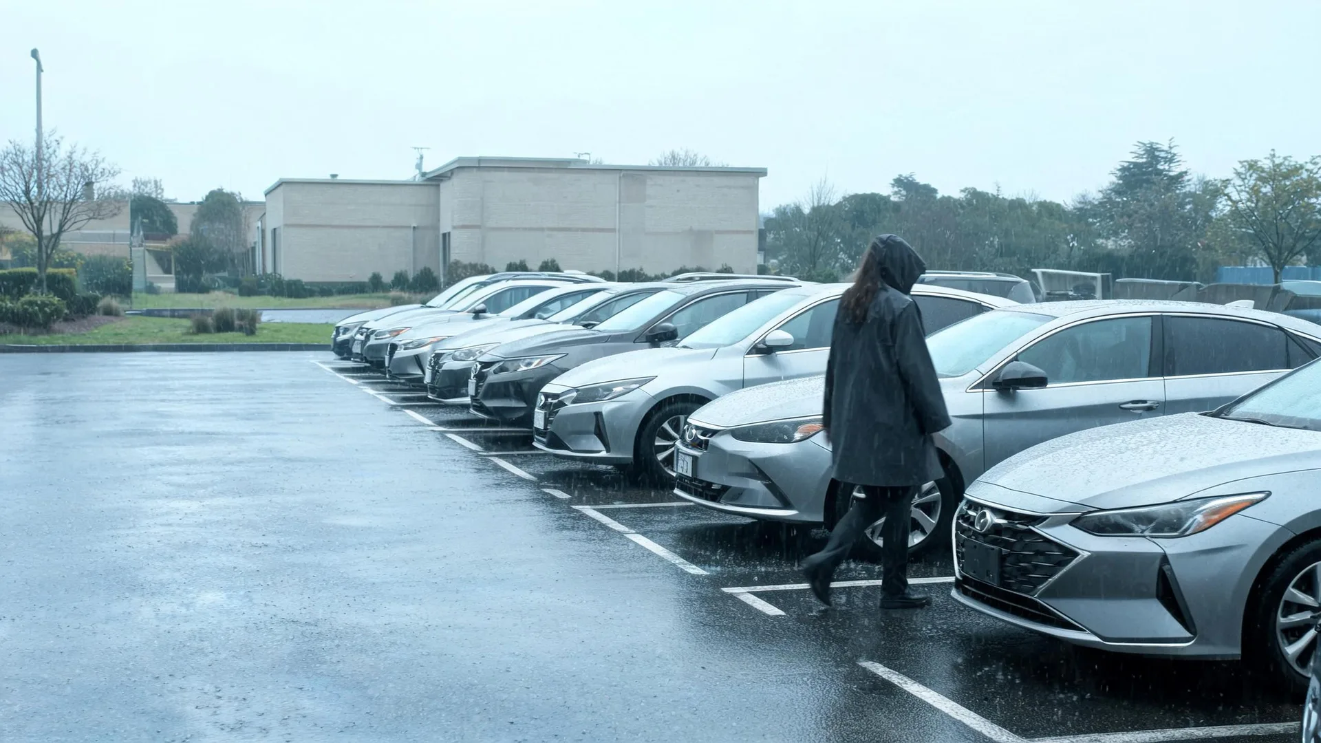 A person in a dark hooded coat walks past a row of parked silver sedans in a wet parking lot under overcast skies, with a low beige building and bare trees in the background.
