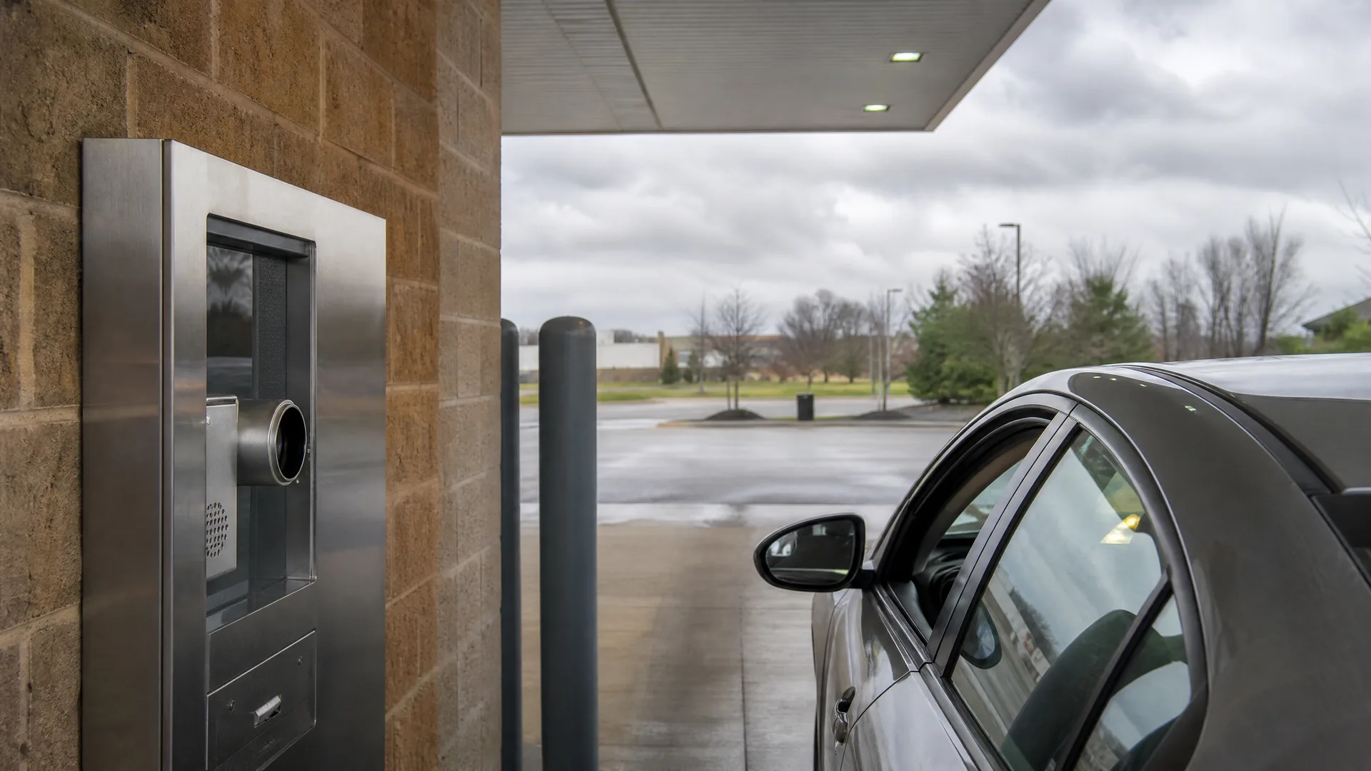 A silver car pulled up under a canopy beside a stainless steel drive-through terminal with a tube carrier mounted on a tan block wall, with wet pavement and bare trees under an overcast sky in the background.