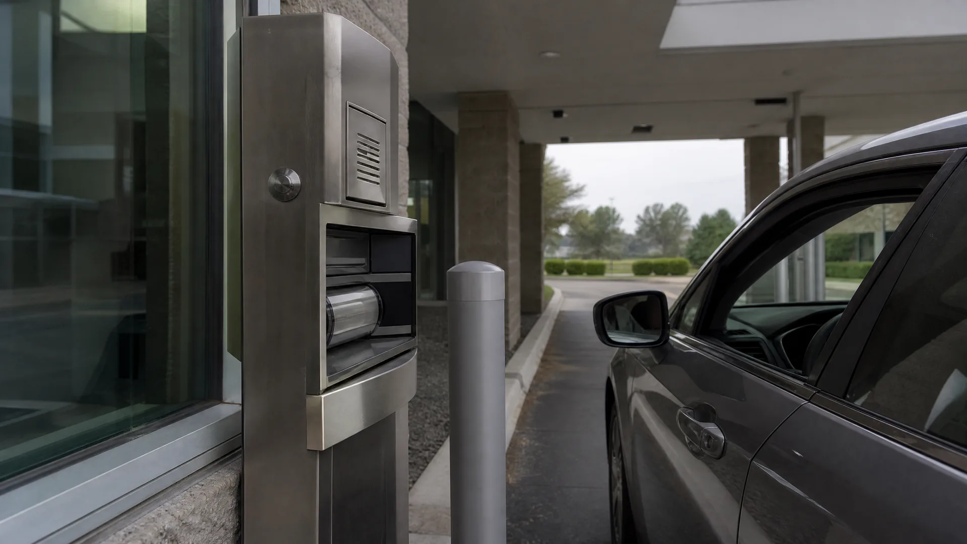 A stainless steel drive-through service terminal with a speaker grille and transaction drawer stands beside a glass building wall, with a gray car stopped at the lane and a metal bollard between them.