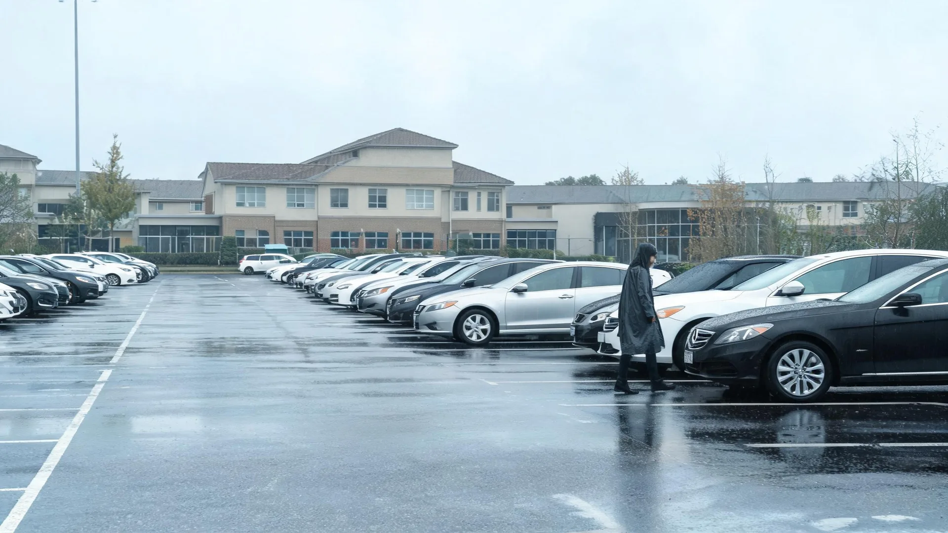 A person in a dark hooded raincoat walks between rows of parked cars on a wet parking lot under an overcast sky, with a two-story light-colored building and bare trees in the background.