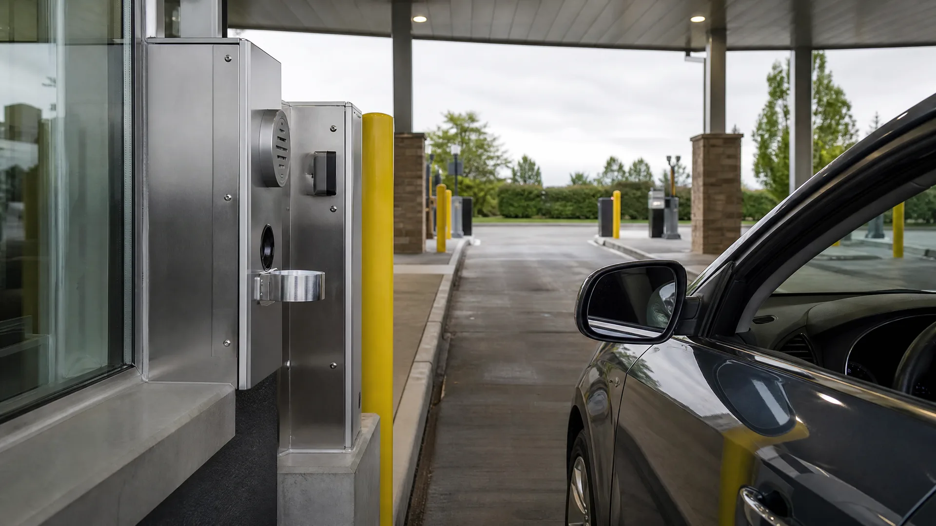 A dark car stops beside a stainless steel drive-through terminal with a pneumatic tube canister under a canopy, with yellow bollards and green hedges in the background.