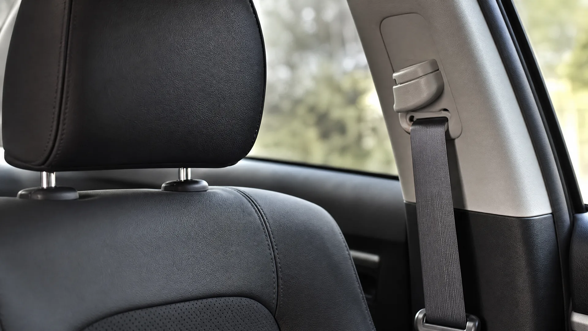 Car interior close-up showing a black leather seat with headrest and a seat belt running through a height adjuster on the pillar, with blurred green foliage visible through the window.