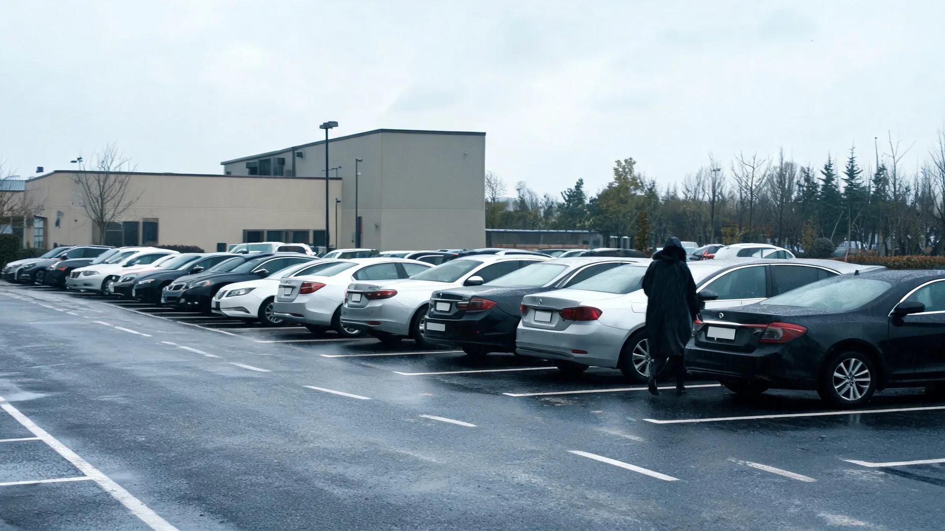 A person in a dark hooded coat walks between rows of parked cars in a wet parking lot under an overcast sky, with a beige flat-roofed building and bare trees behind.