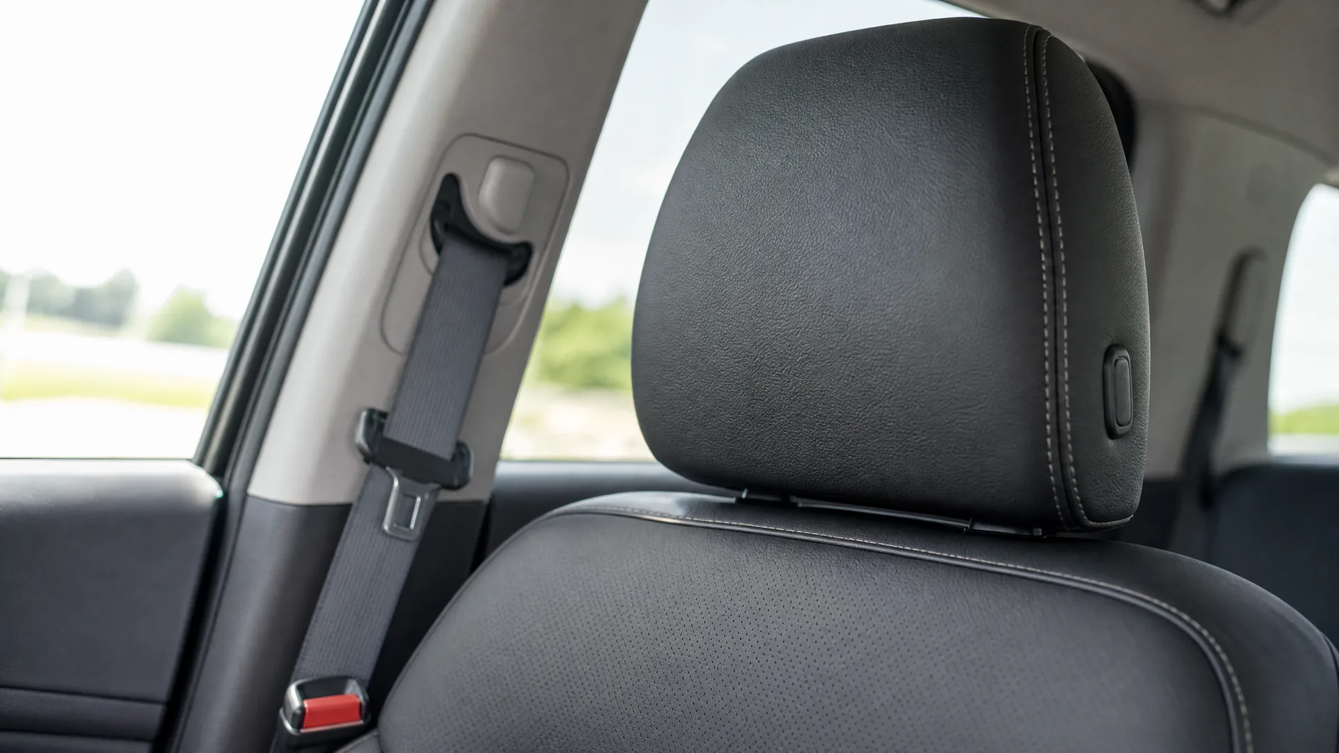 Close-up of a black leather car front seat headrest and seatback, with a seat belt and buckle beside the window showing blurred green trees outside.