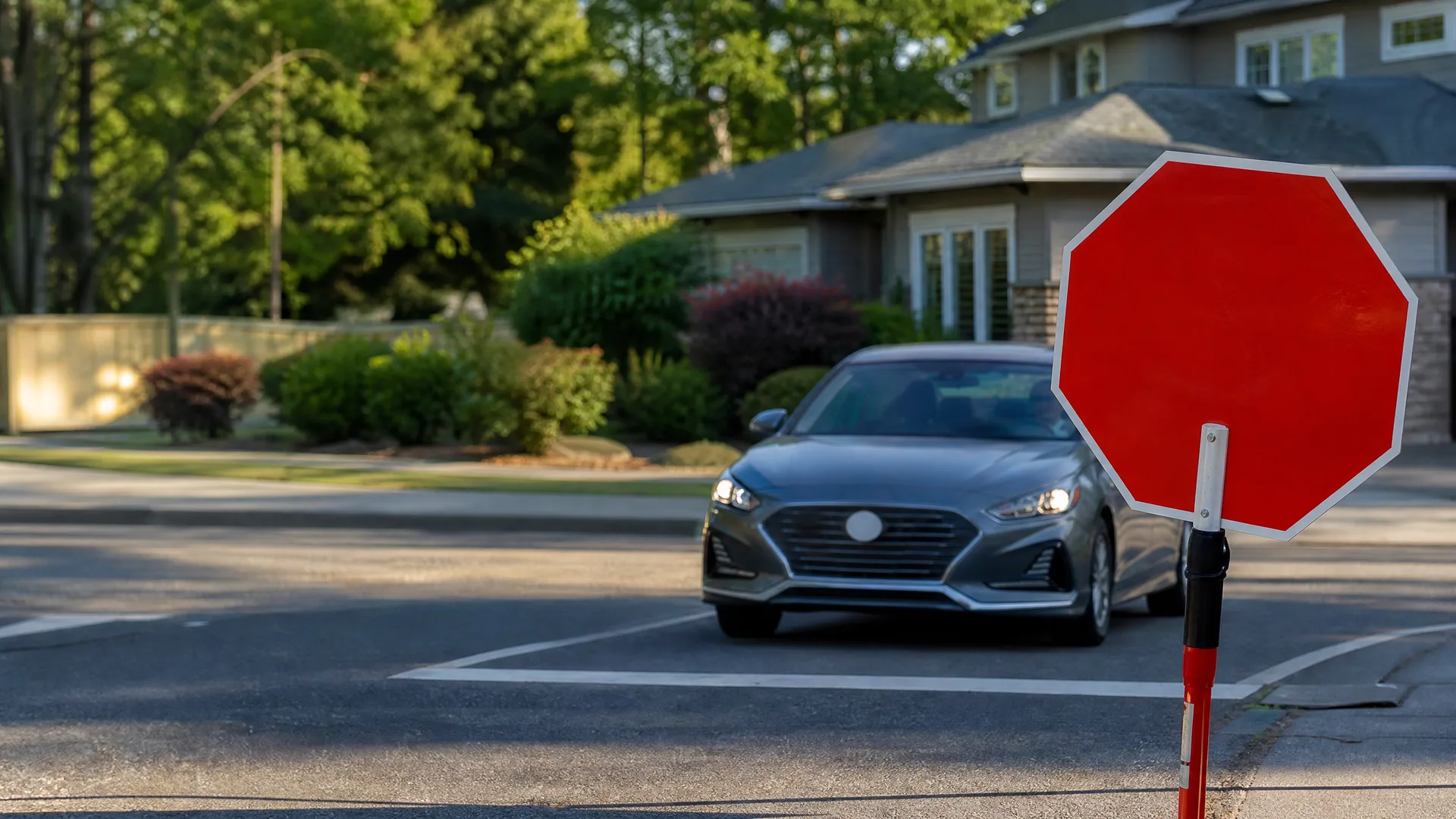 A silver sedan stops at a crosswalk on a suburban street, with a blank red octagonal handheld stop sign held in the right foreground and a house and trees behind.