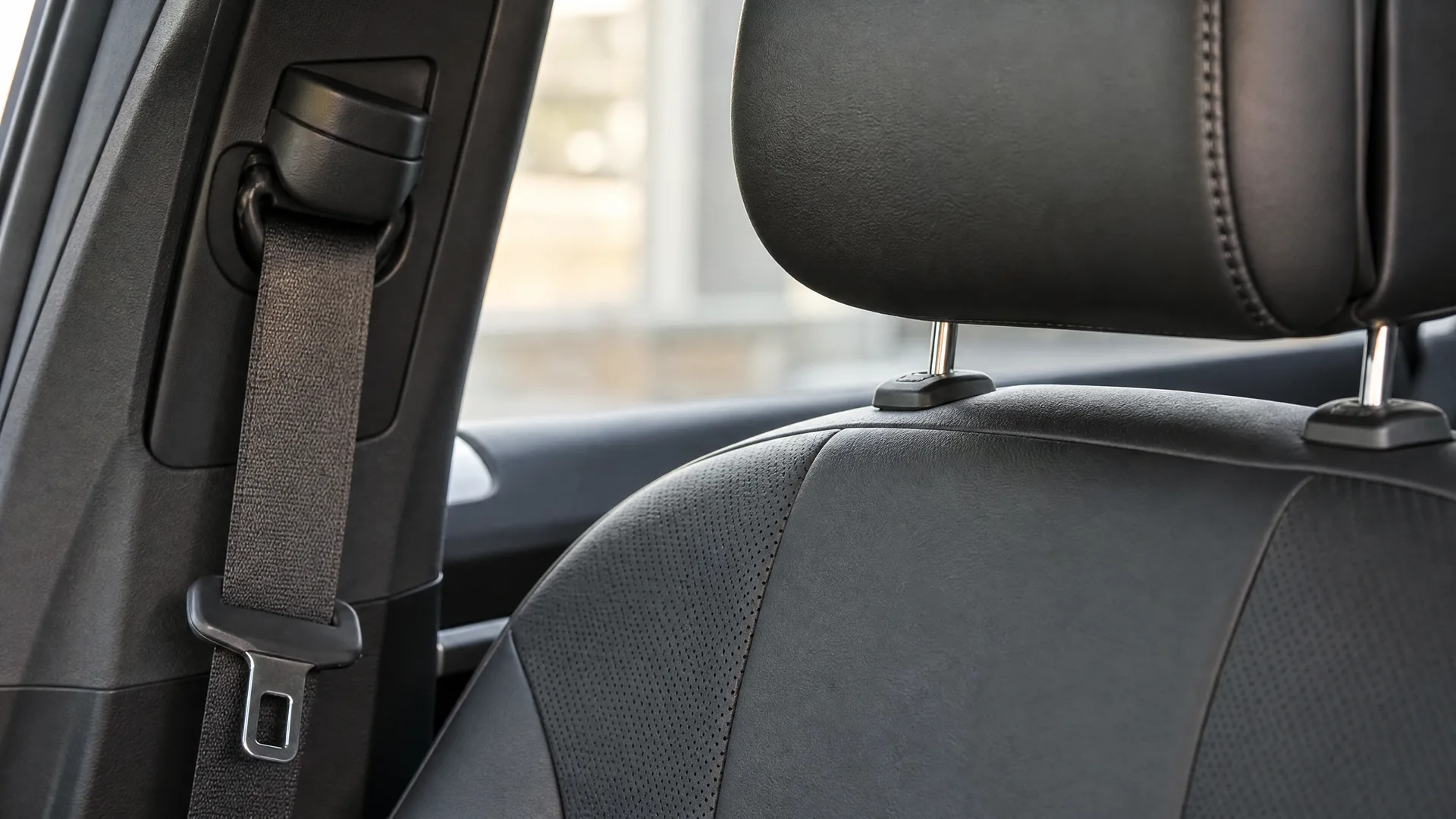 Close-up of a car interior showing a black seat belt against the door pillar and a black perforated leather seat with headrest, with a bright window behind.