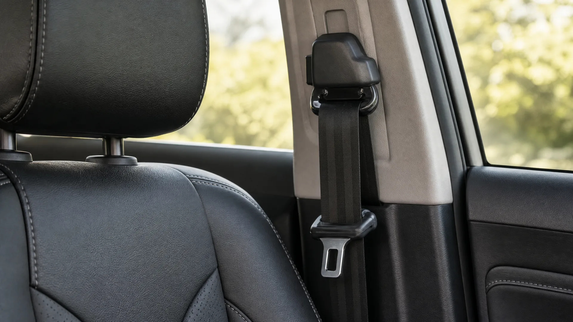 Car interior close-up showing a black leather seat with headrest and a seat belt with metal buckle on the B-pillar, with blurred green foliage through the window.
