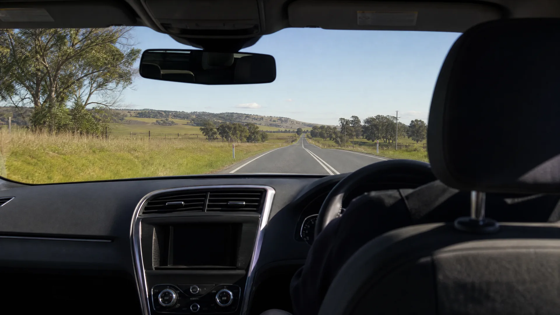 View through a car windscreen from the back seat, showing the dashboard, rear-view mirror and right-hand steering wheel, with a straight two-lane road crossing grassy farmland toward low hills under a clear sky.