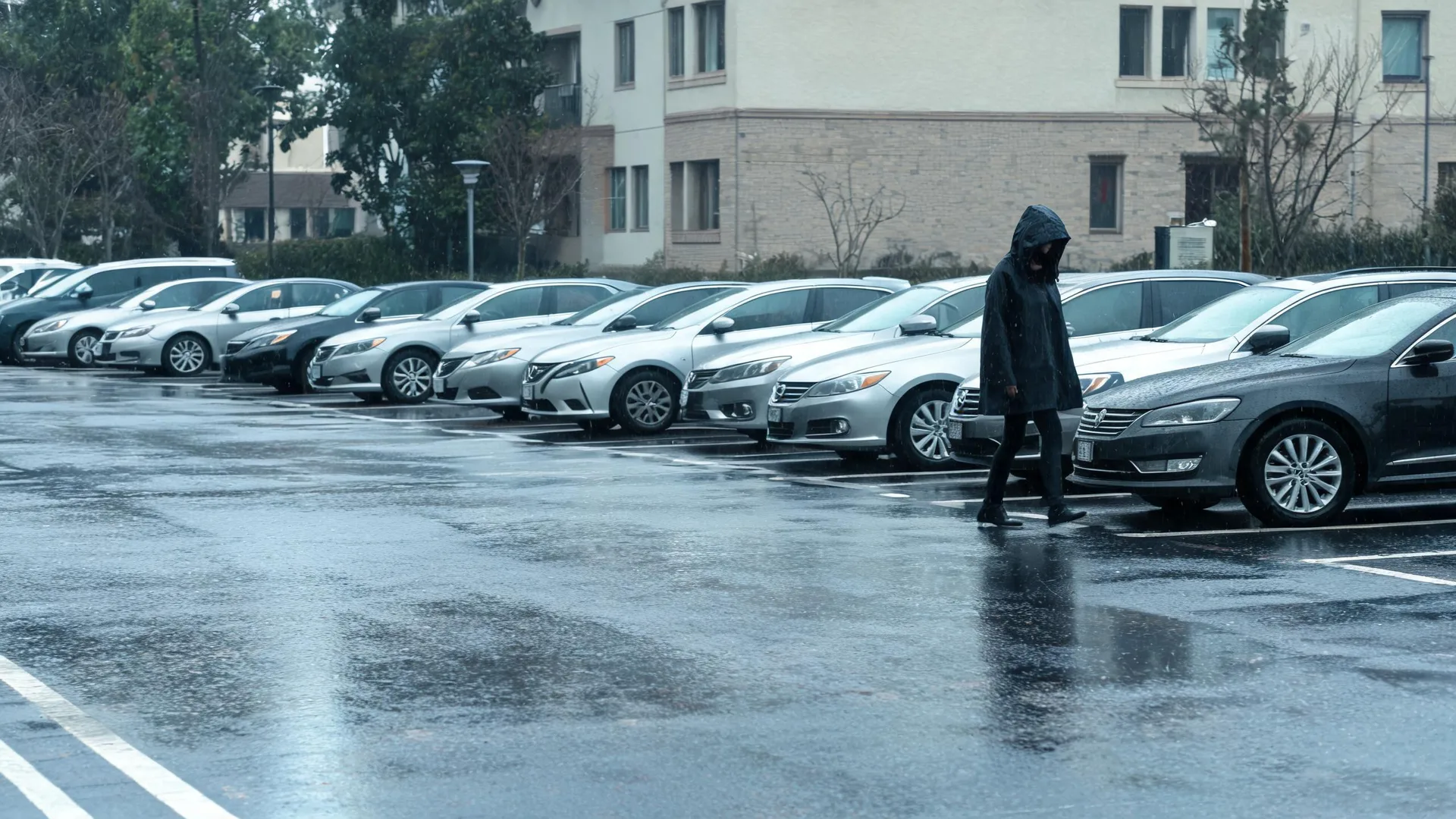 A person in a dark hooded raincoat walks across a wet parking lot past a row of parked cars, with a multi-story building and bare trees in the background.