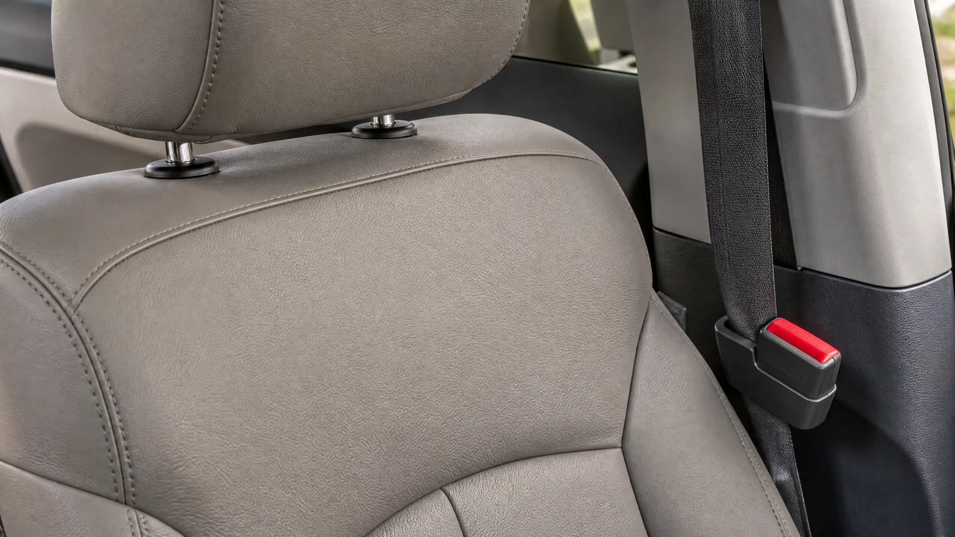 Close-up of a light gray leather vehicle front seat back and headrest, with a black seat belt and red buckle release on the pillar behind it.