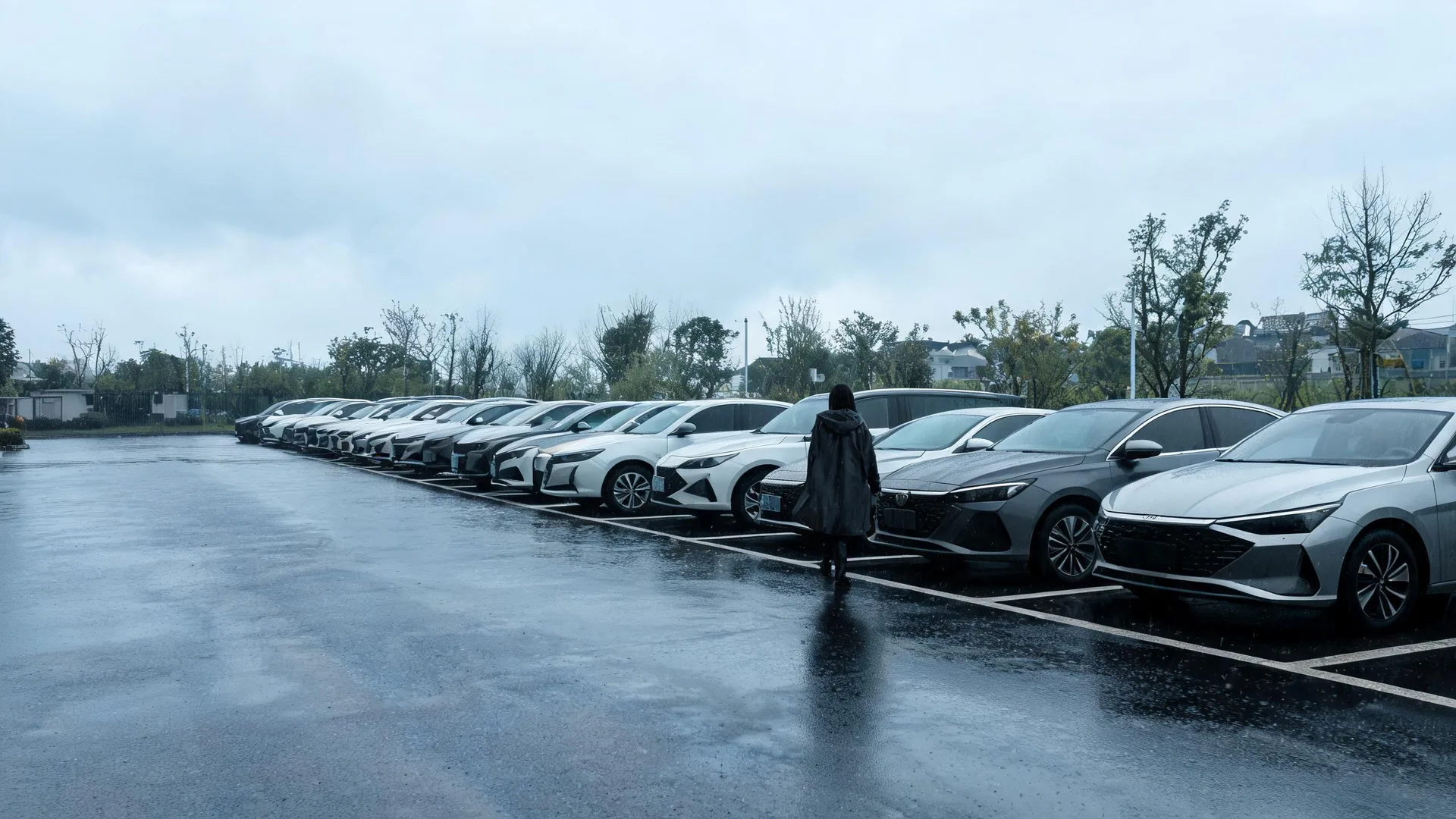 A person in a dark hooded raincoat walks along a wet parking lot lined with a long row of parked cars under an overcast sky, with bare trees and low buildings in the background.