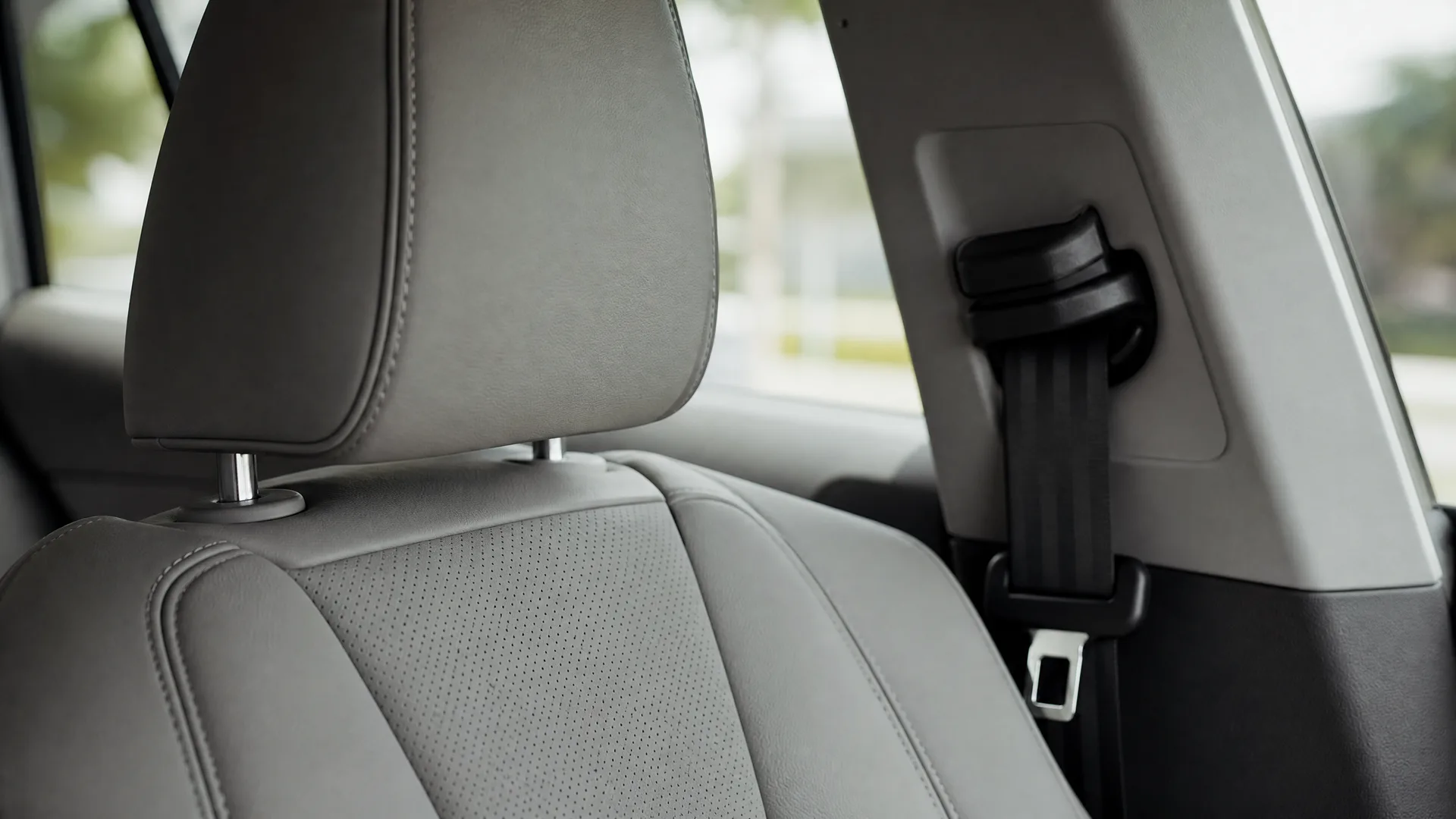 Close-up of a light gray leather car front seat with headrest, and a black seat belt with buckle on the door pillar, with blurred greenery visible through the window.