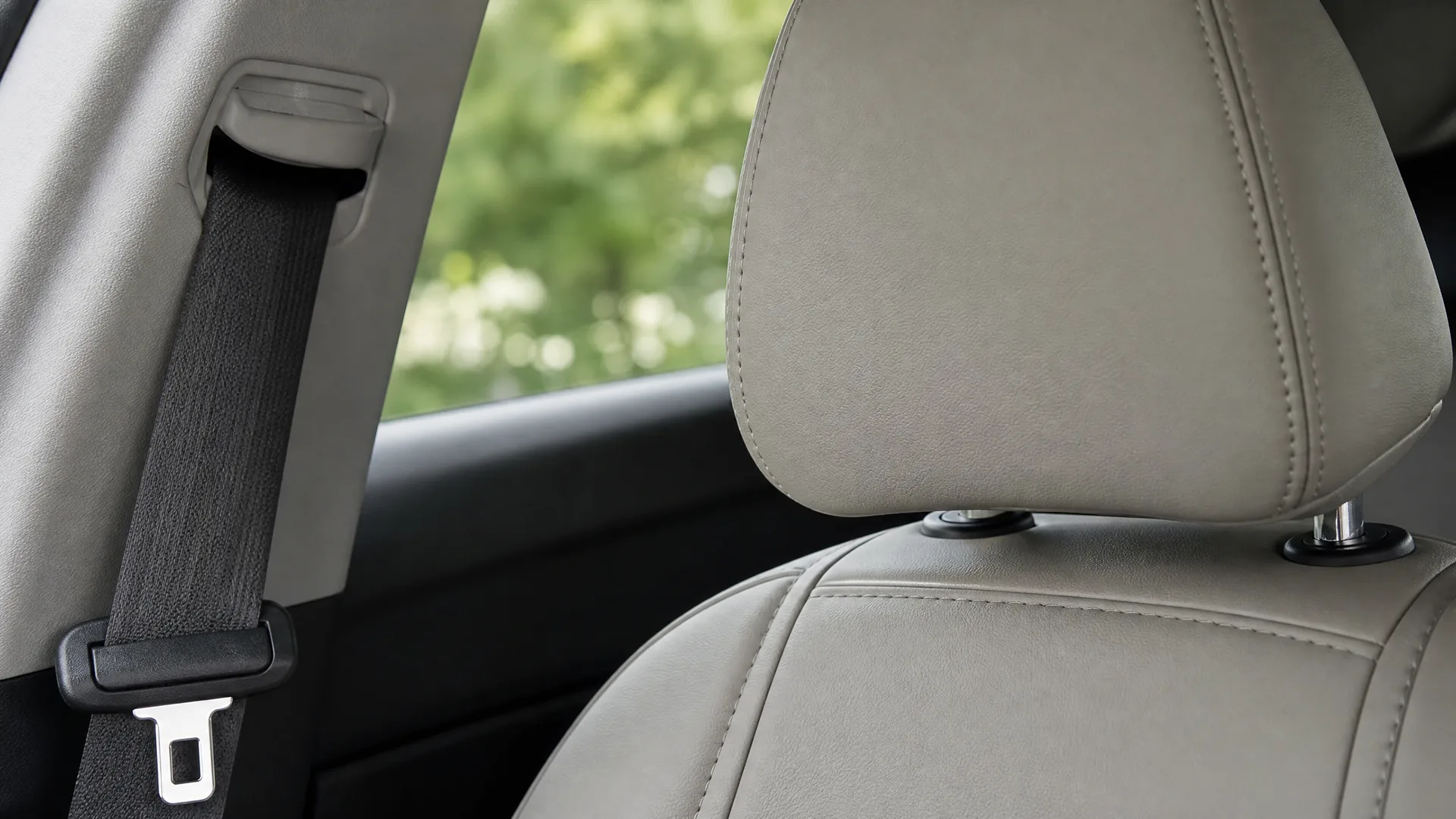Car interior showing a light grey leather seat back and headrest with a black seat belt and buckle beside a window with blurred green foliage outside.
