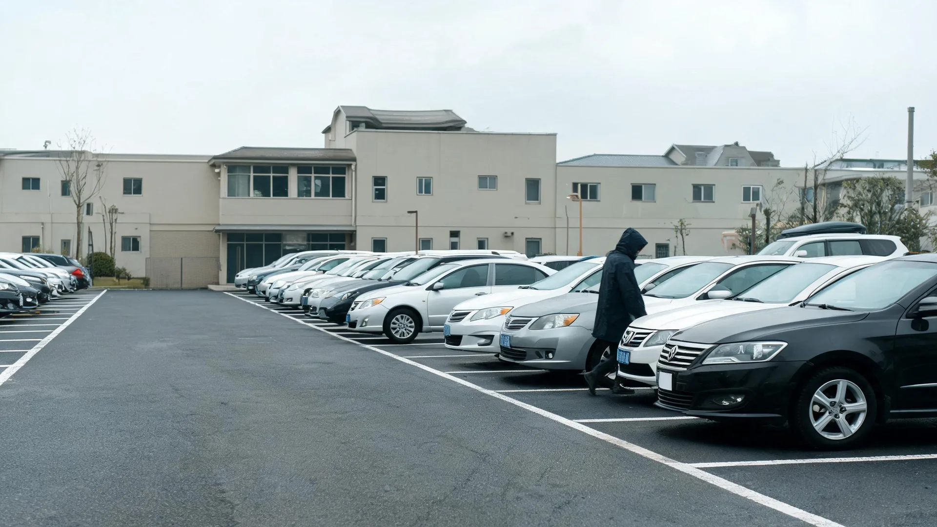 A person in a dark hooded coat walks between rows of parked cars in an outdoor asphalt parking lot in front of a two-story light-colored building under an overcast sky.