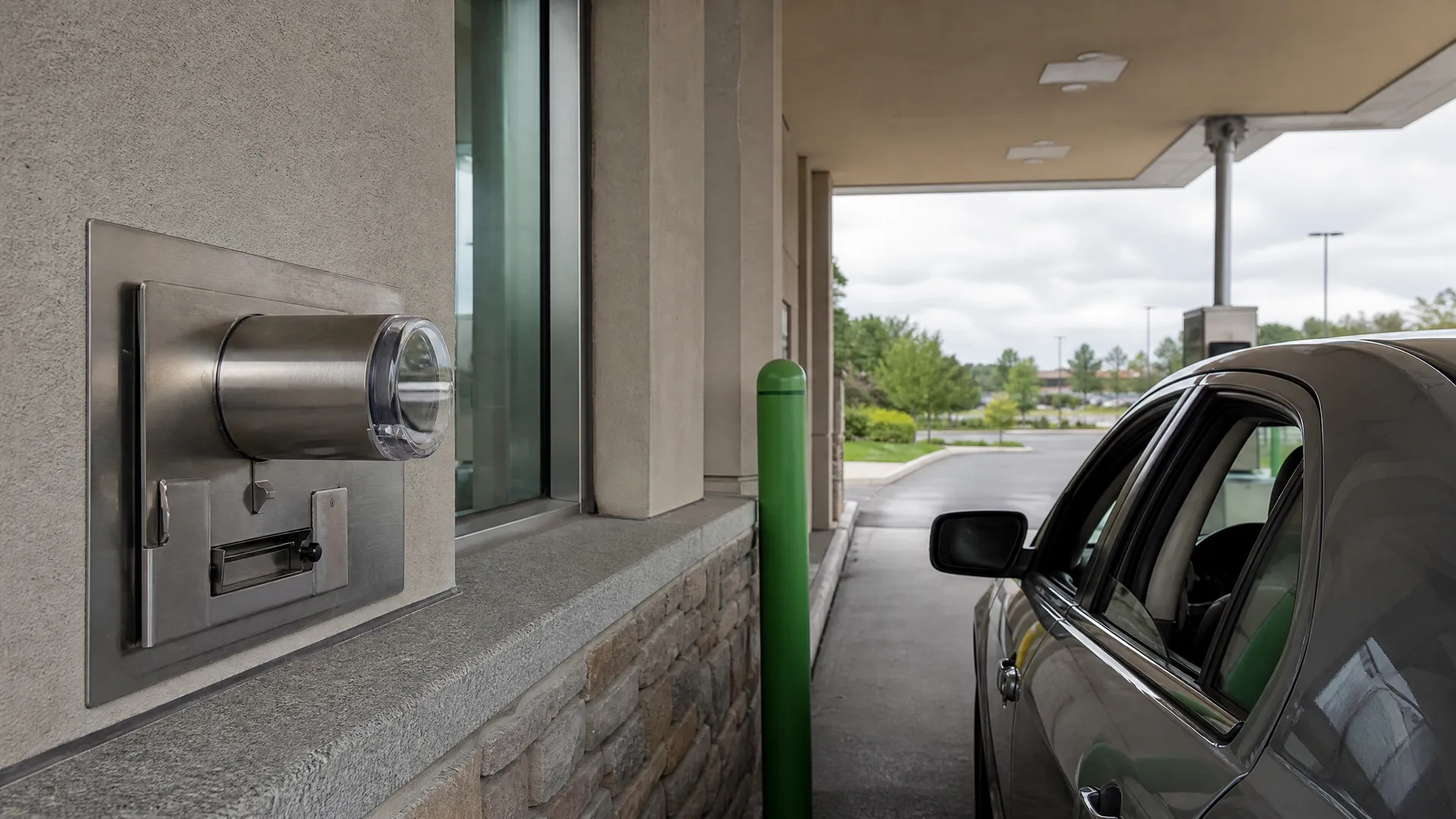 A stainless steel drive-up teller window with pneumatic tube and drawer set in a stucco and stone wall, with a dark sedan waiting in the lane beneath a canopy.