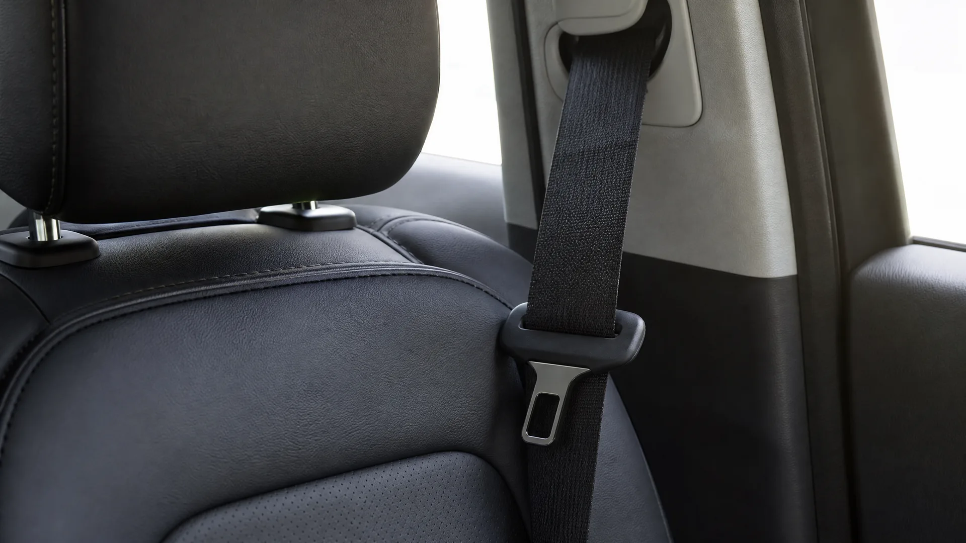 Close-up of a black seat belt with metal latch plate beside a dark leather car seat and headrest, with a bright window behind.
