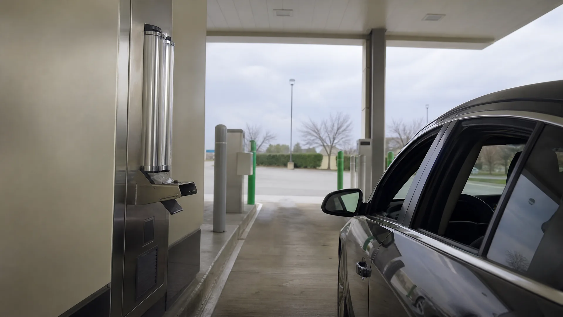 A dark sedan stopped beside a stainless steel drive-through terminal under a covered canopy, with bare trees and an empty parking lot in the background under an overcast sky.