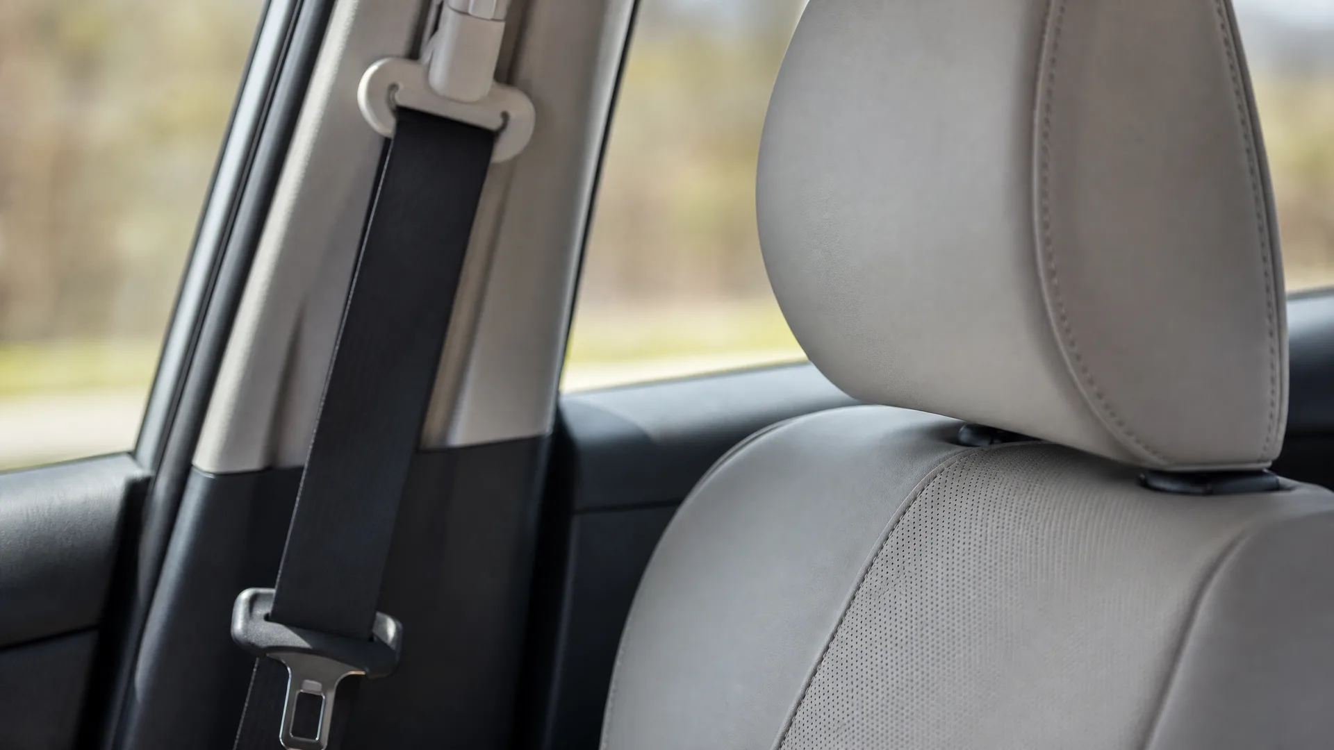 Close-up of a car interior showing a black seat belt along the door pillar and a light gray leather seat with headrest, with blurred greenery visible through the window.