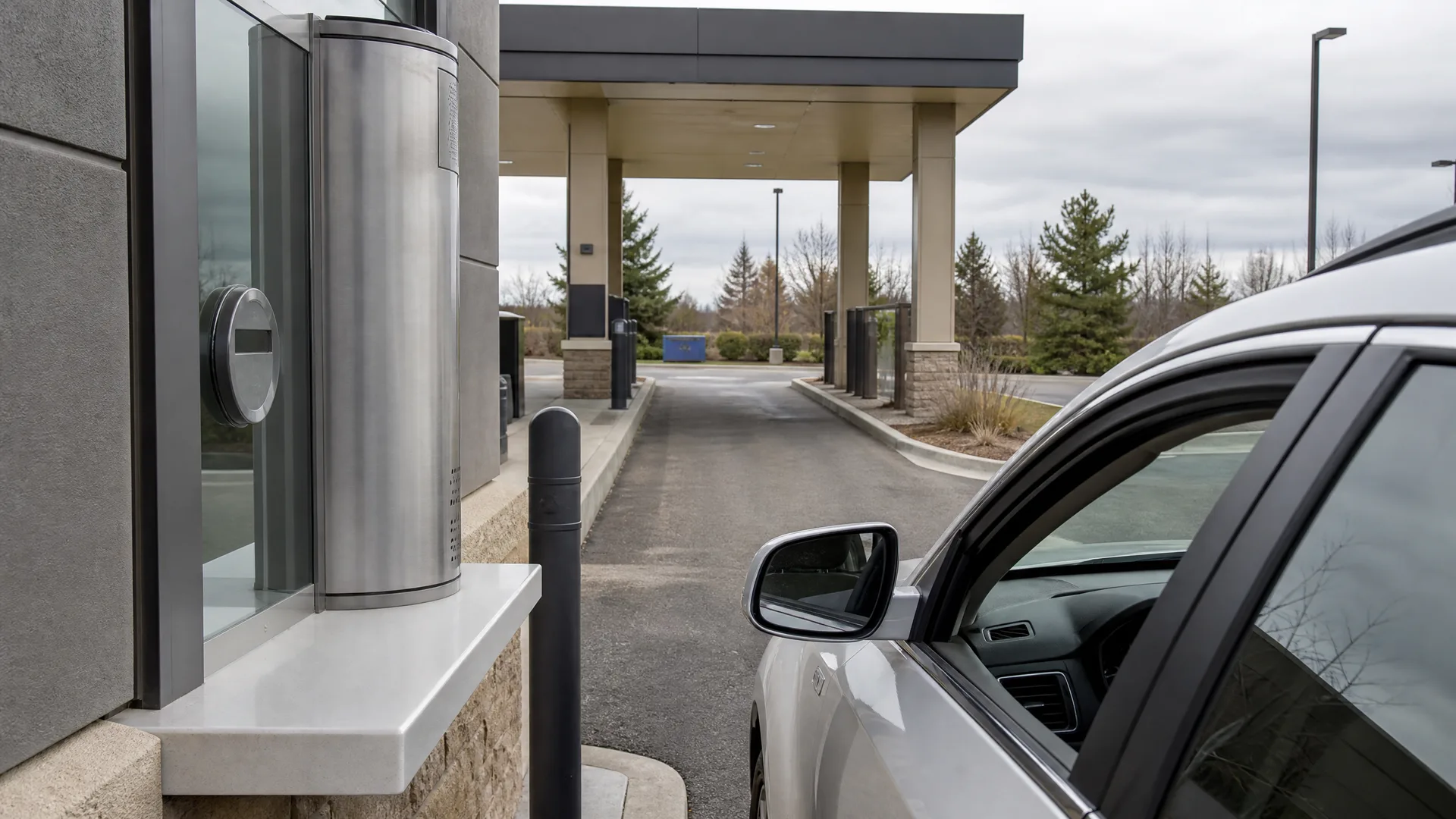 A silver car parked at a drive-up window with a stainless steel transaction unit on a stone ledge, with a covered drive-through canopy and evergreen trees under an overcast sky in the background.