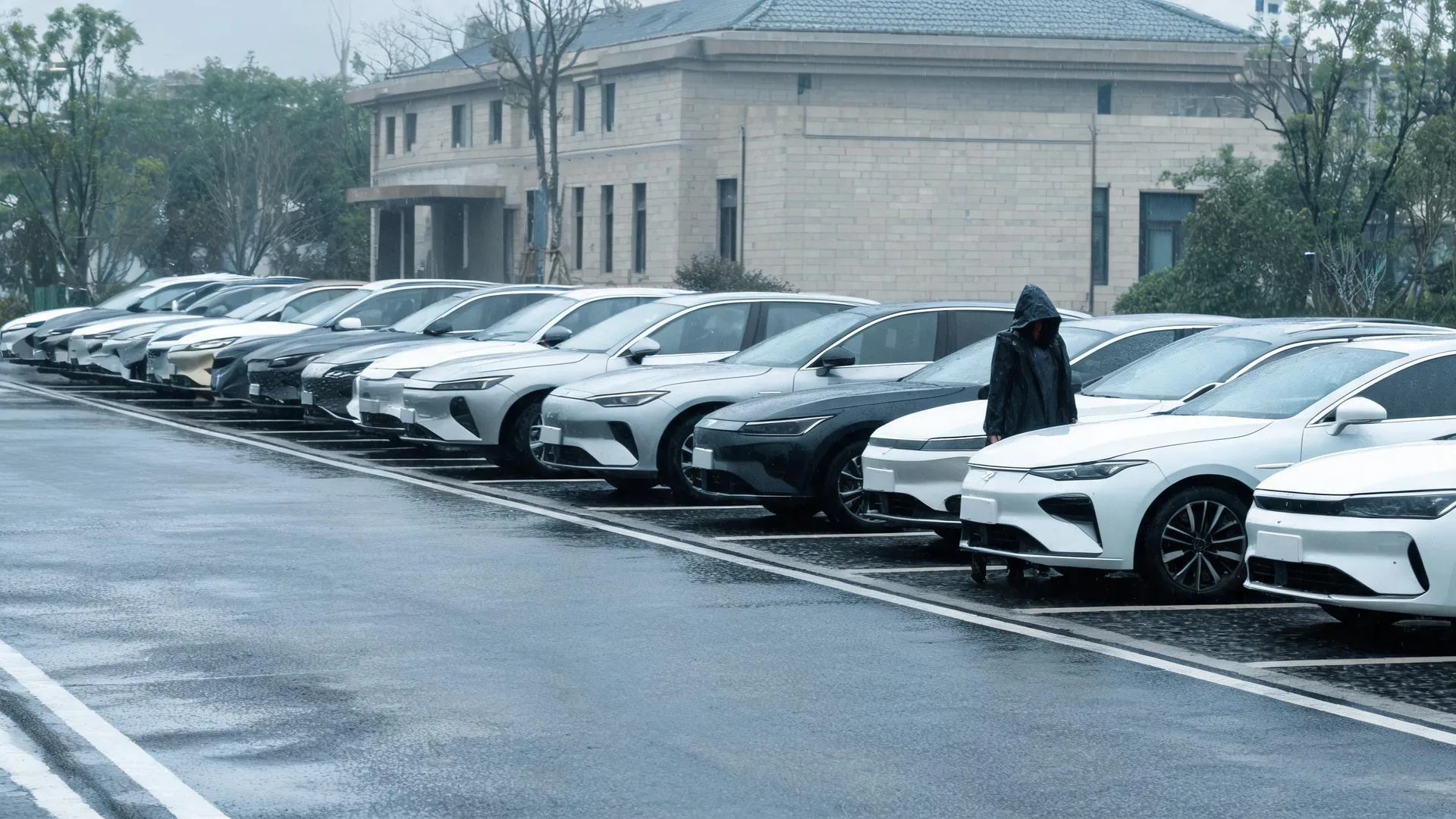 A long row of white, grey and black cars parked diagonally on wet pavement, with a person in a dark hooded raincoat standing between two of them and a stone-clad two-storey building and bare trees behind.