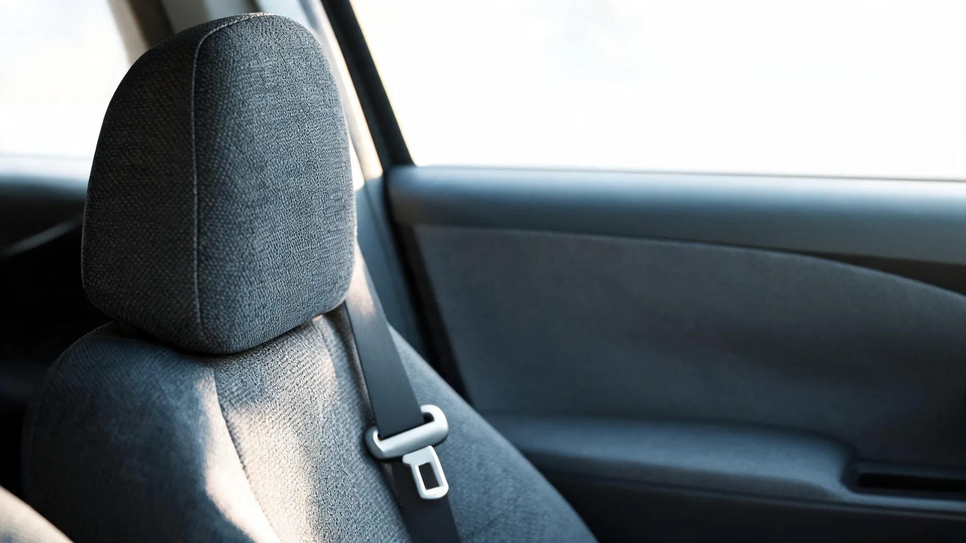 Close-up of a gray fabric car seat with headrest and a seat belt buckle tongue, beside a car door panel and bright window.