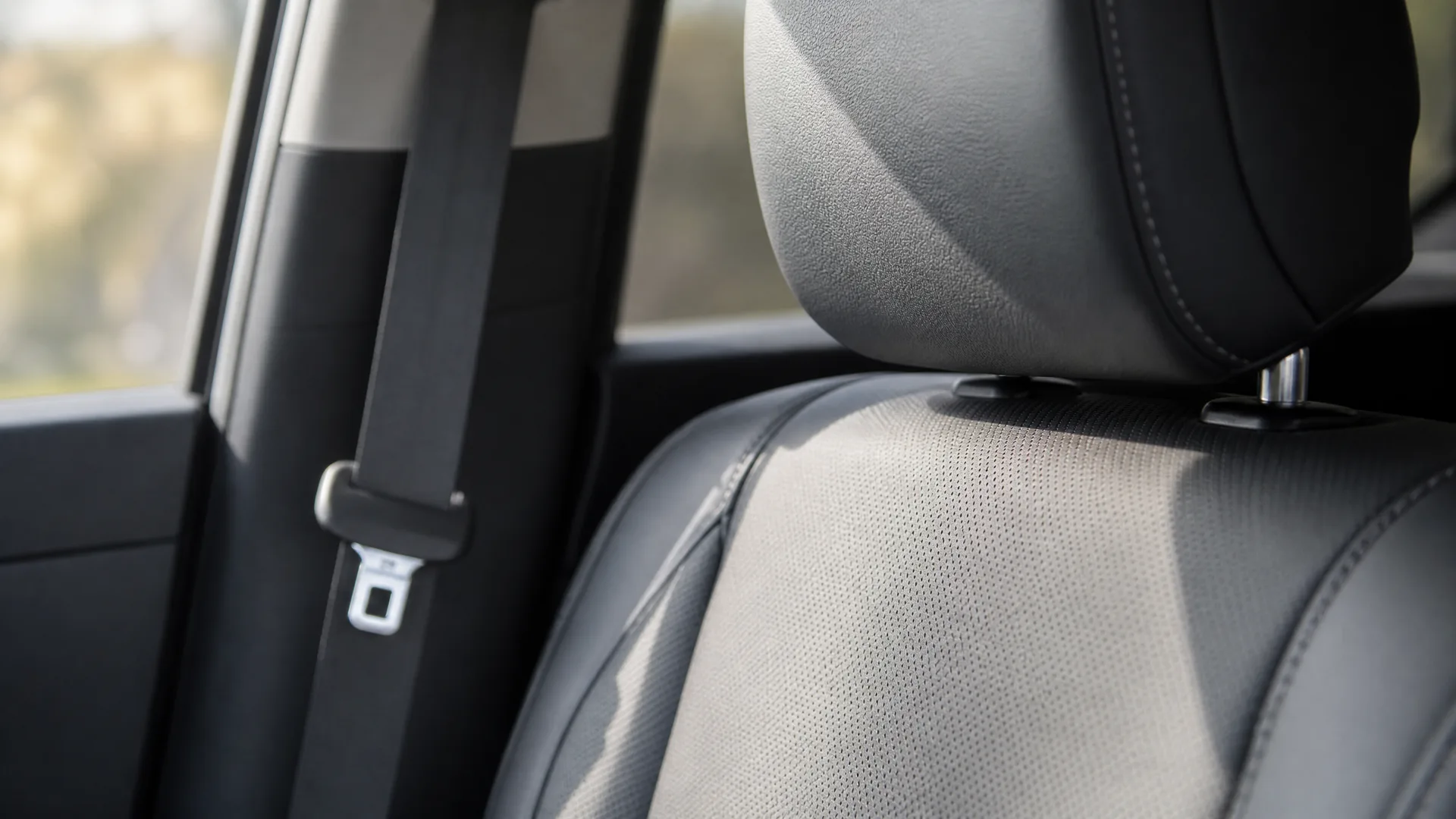 Close-up of a car's front seat with perforated dark upholstery, headrest, and a seat belt running along the door pillar beside a bright window.