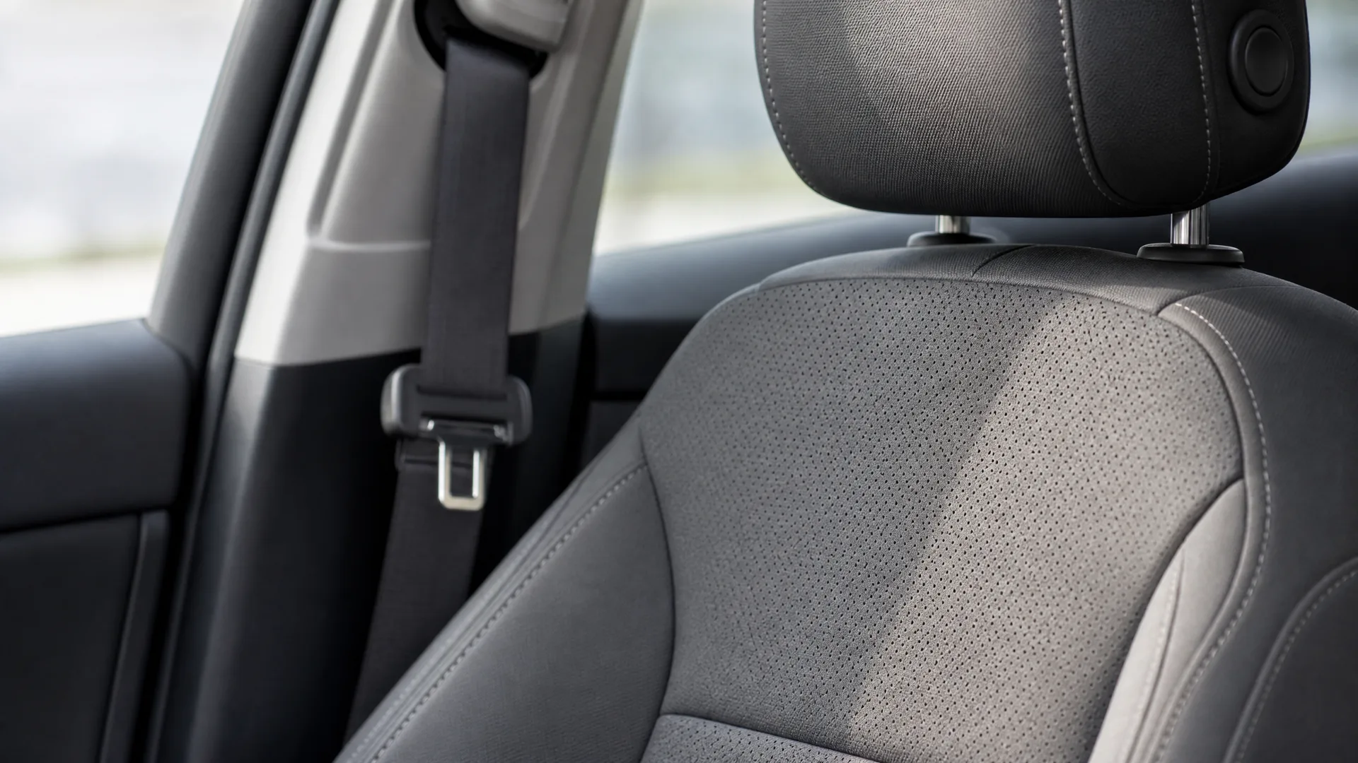 Close-up of a car's front seat with perforated dark upholstery, adjustable headrest, and a black seat belt against the door pillar, with a blurred window view.