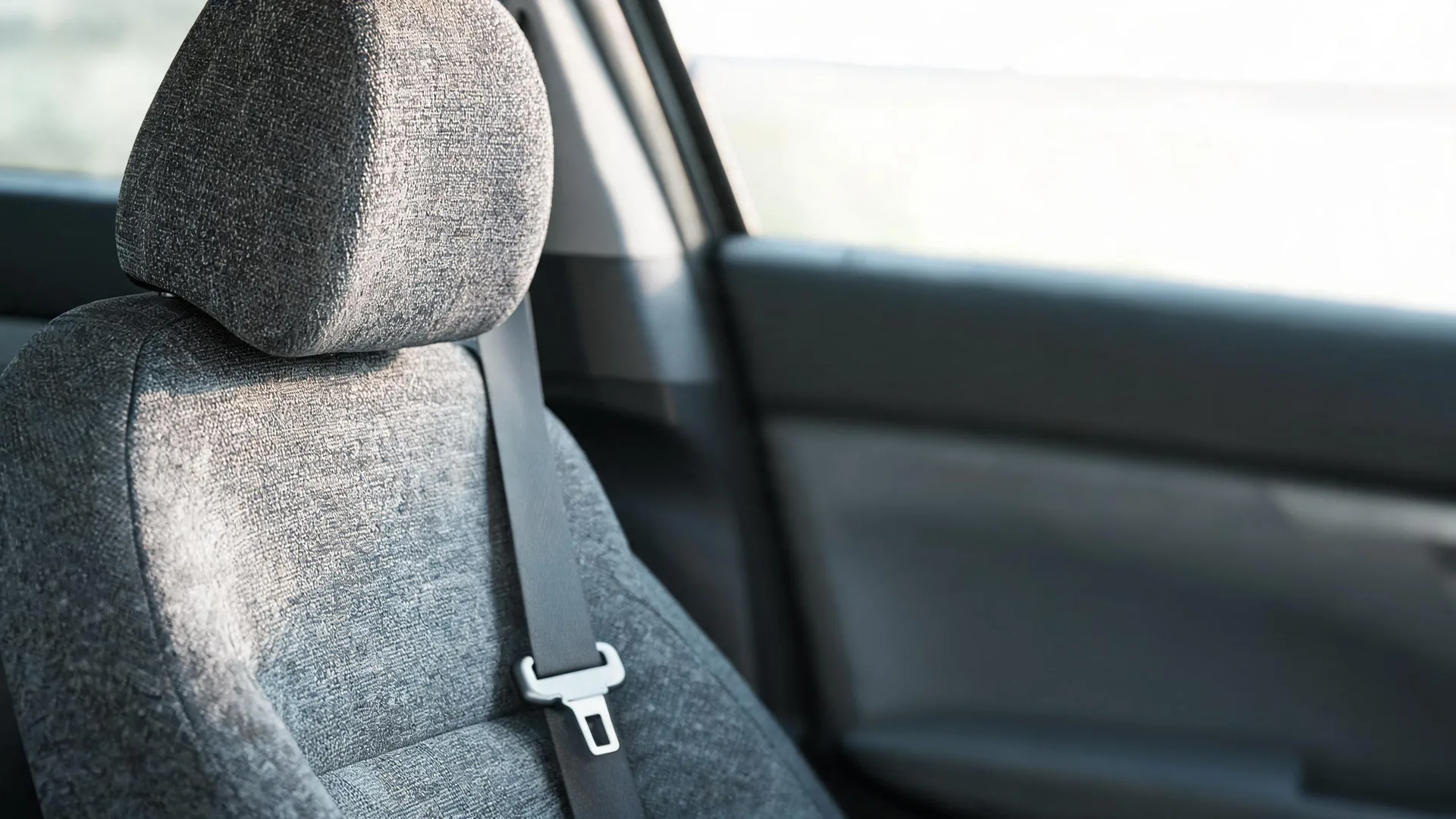 Close-up of a gray fabric car seat with headrest and a seat belt with metal buckle, beside a window and door panel inside a vehicle.