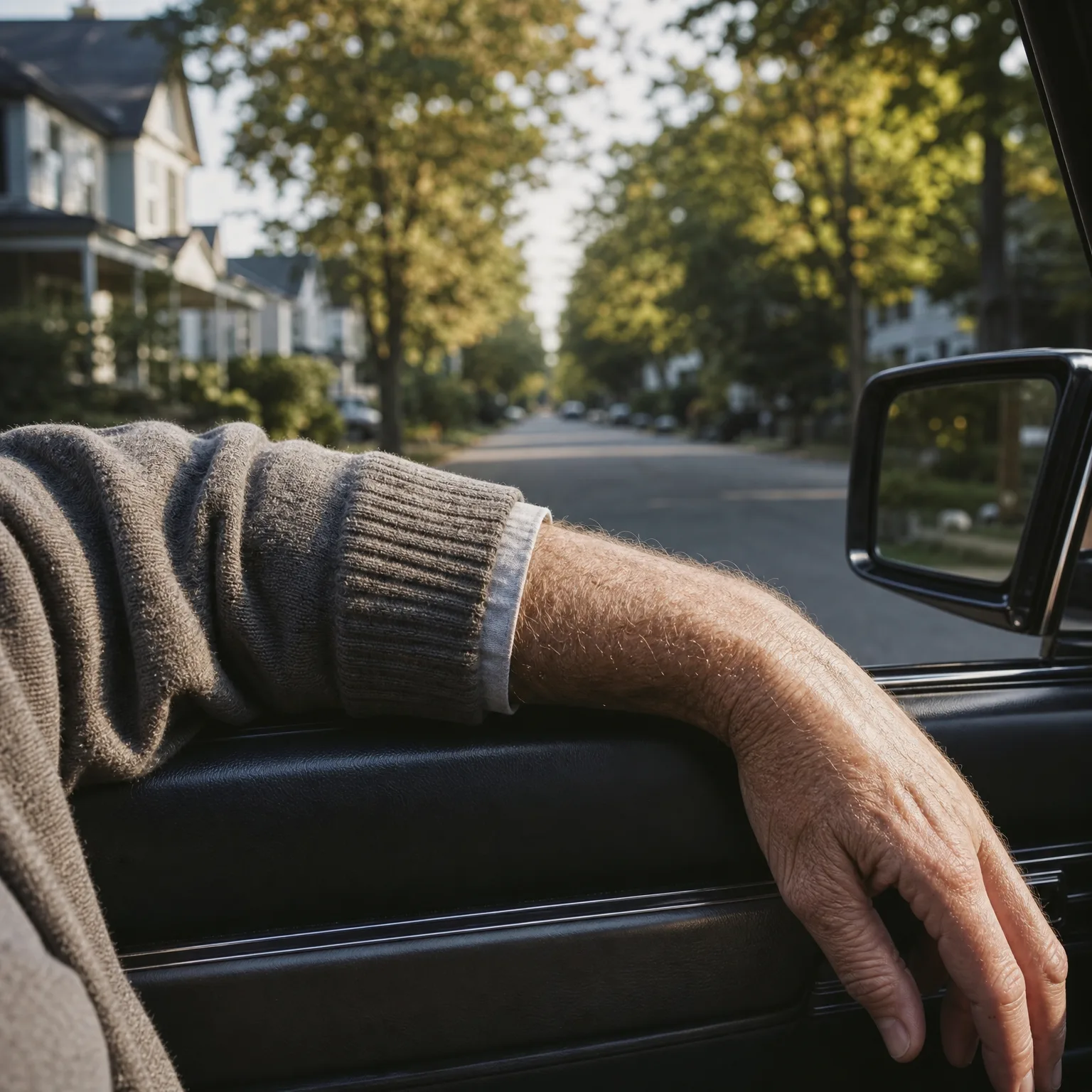 Close-up of a person's arm in a knit sweater resting on the open window sill of a car door, with a tree-lined residential street and side mirror visible beyond.