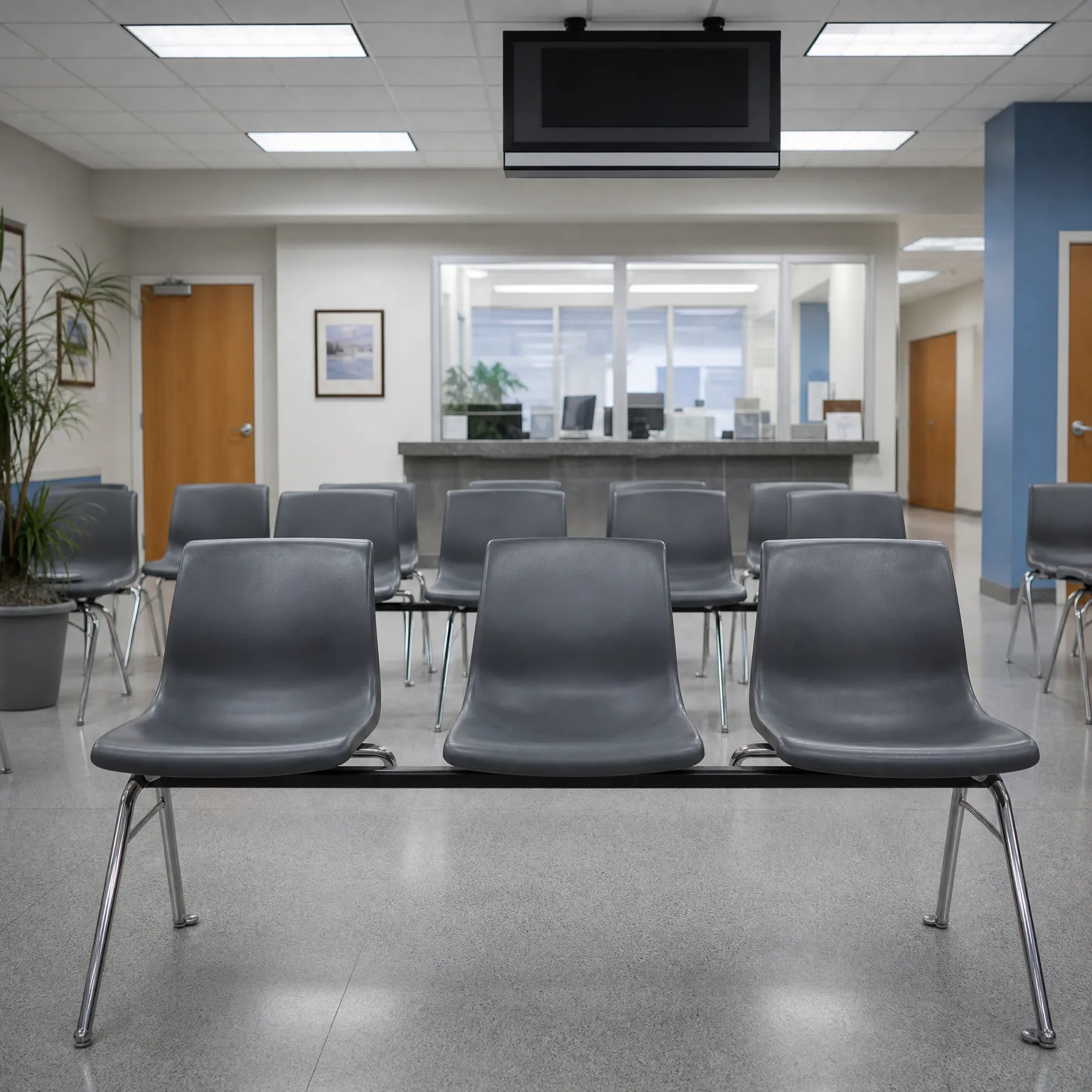 An empty indoor waiting area with rows of gray molded plastic beam chairs on speckled tile flooring, a reception counter with a glass window, a wall-mounted flat screen, and a potted palm at left.