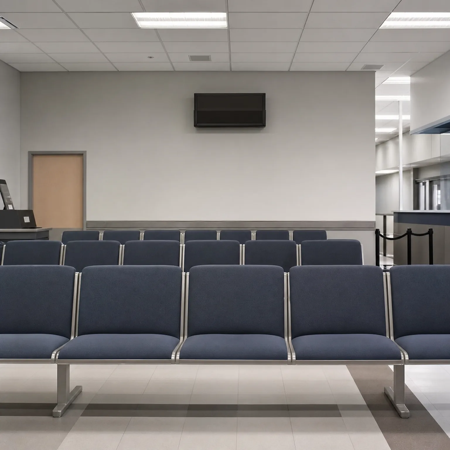 Empty waiting area with rows of blue upholstered beam seating on metal frames, a wall-mounted flat screen, a wooden door, and a tiled floor under a suspended ceiling with fluorescent panels.