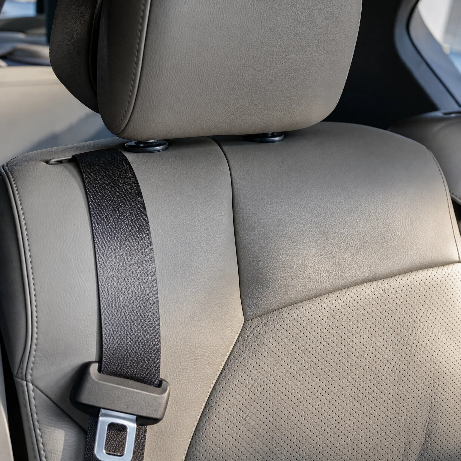 Close-up of a beige leather car seat back with headrest posts, perforated upholstery panel, and a black seat belt strap with metal latch plate.
