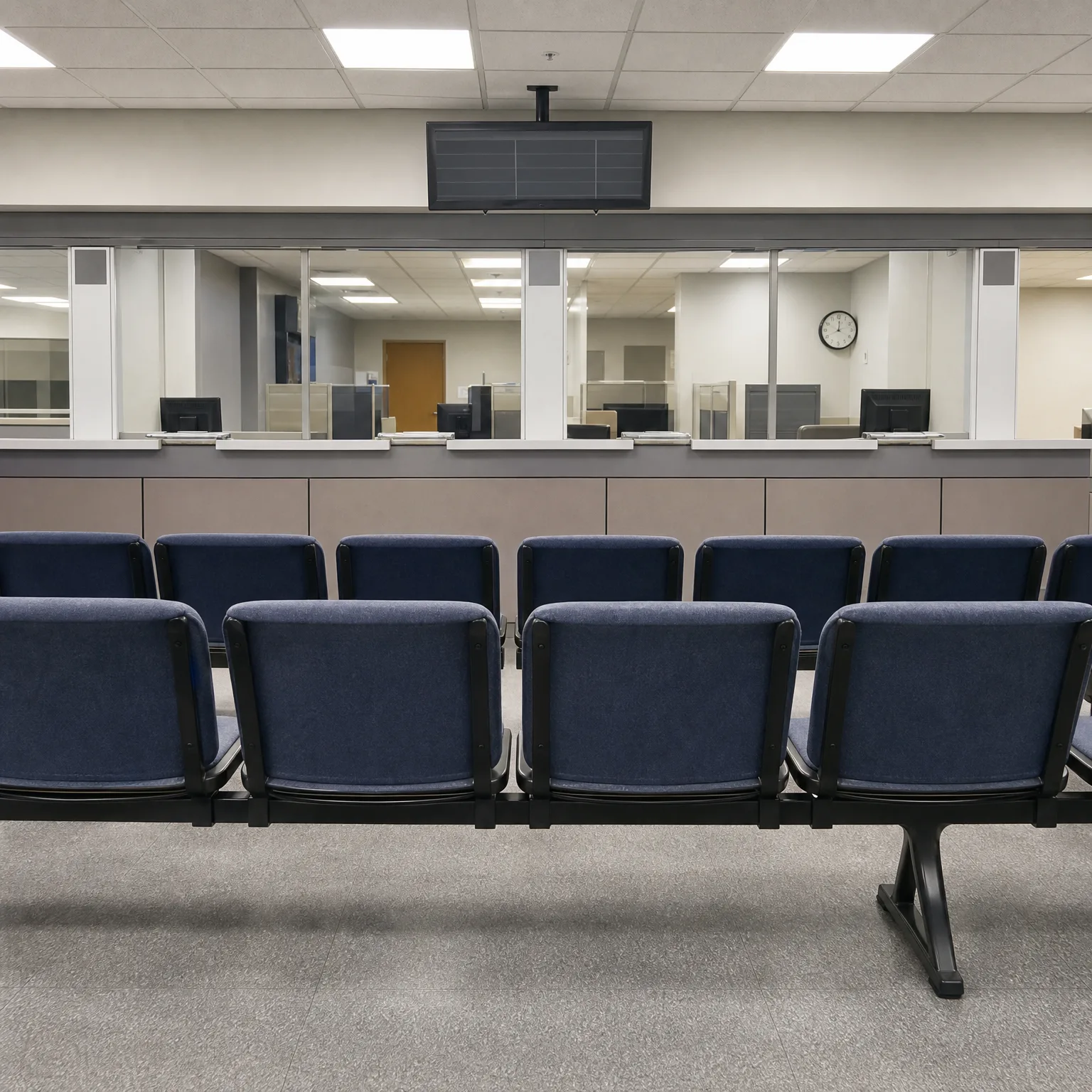 Empty waiting area with two rows of blue upholstered beam seating facing a service counter with glass partitions, a ceiling-mounted monitor, and a wall clock in the office behind.