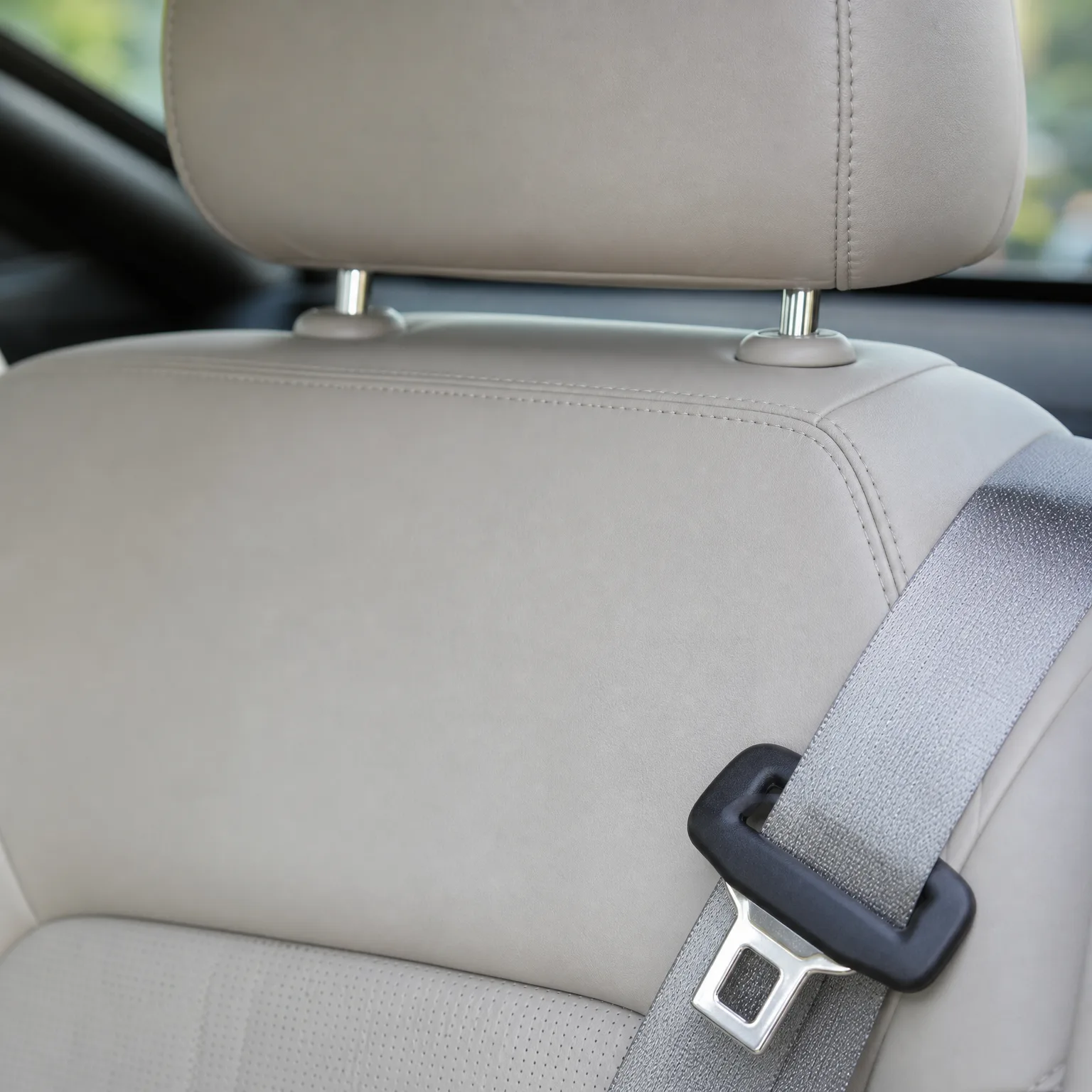 Close-up of a beige leather car seat back and headrest with a gray seat belt and its metal latch plate with black guide on the right.