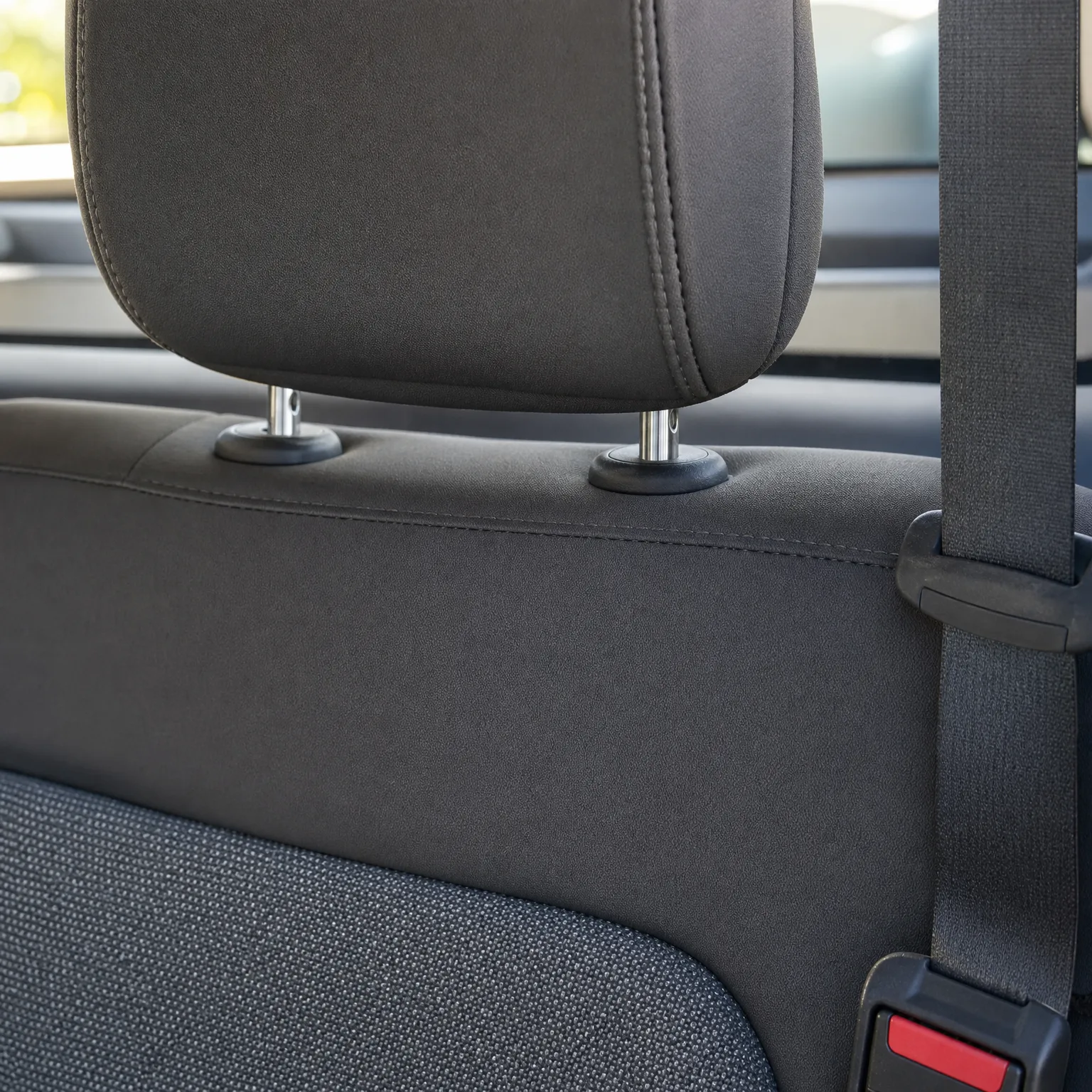 Close-up of a dark grey fabric car seat back with an adjustable headrest on metal posts, a seat belt strap running down the right side, and a red-buttoned buckle at lower right.