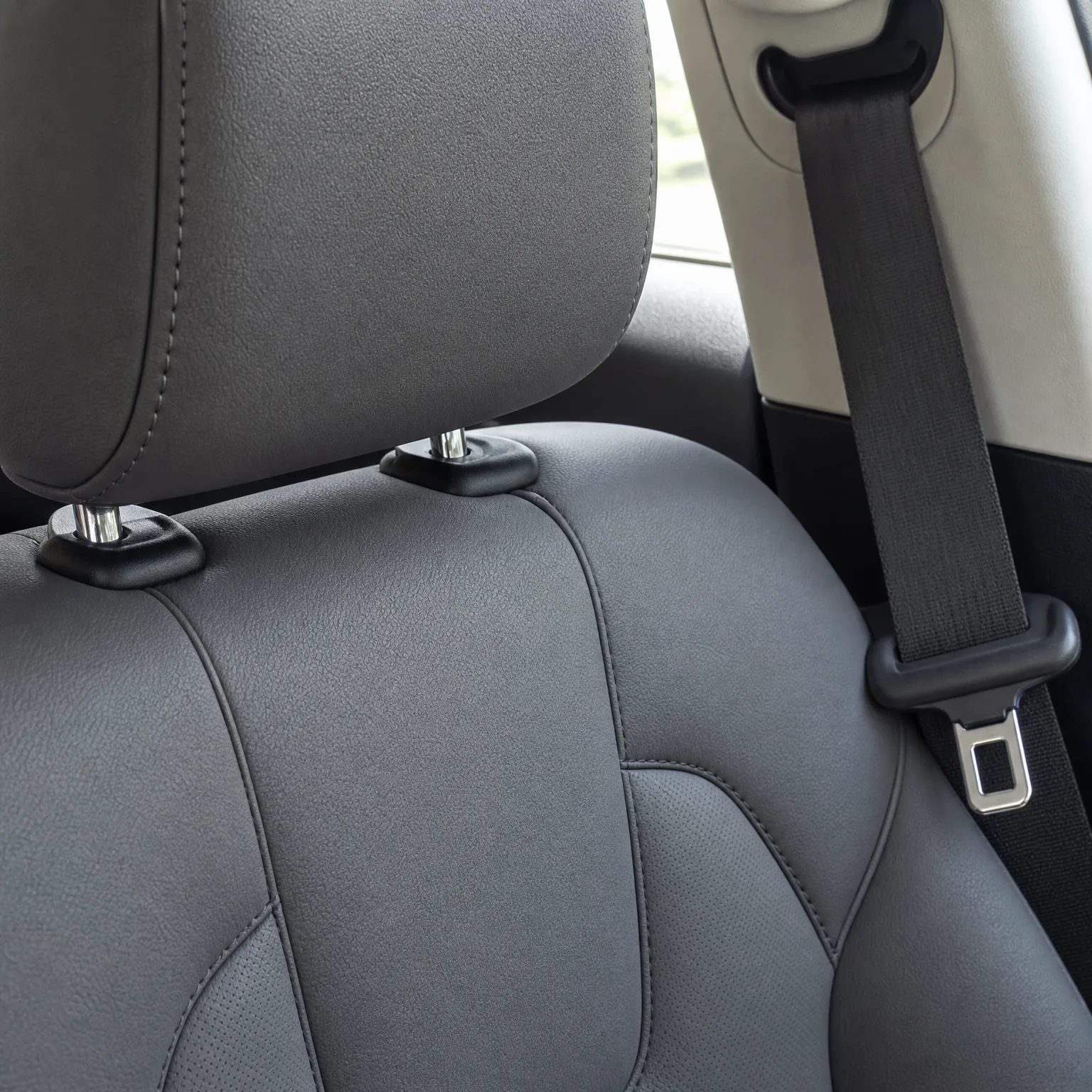 Close-up of a dark gray leather car seat with headrest posts and a seat belt with metal buckle tongue beside the window pillar.