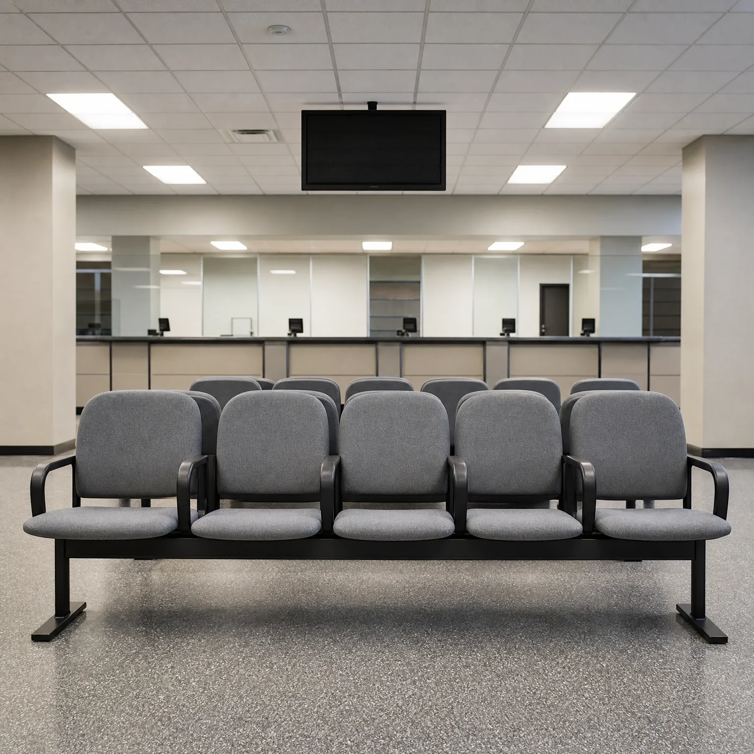 Empty waiting area with two rows of gray beam-mounted chairs facing a long service counter, beneath a ceiling-mounted flat-screen monitor and drop-ceiling light panels.