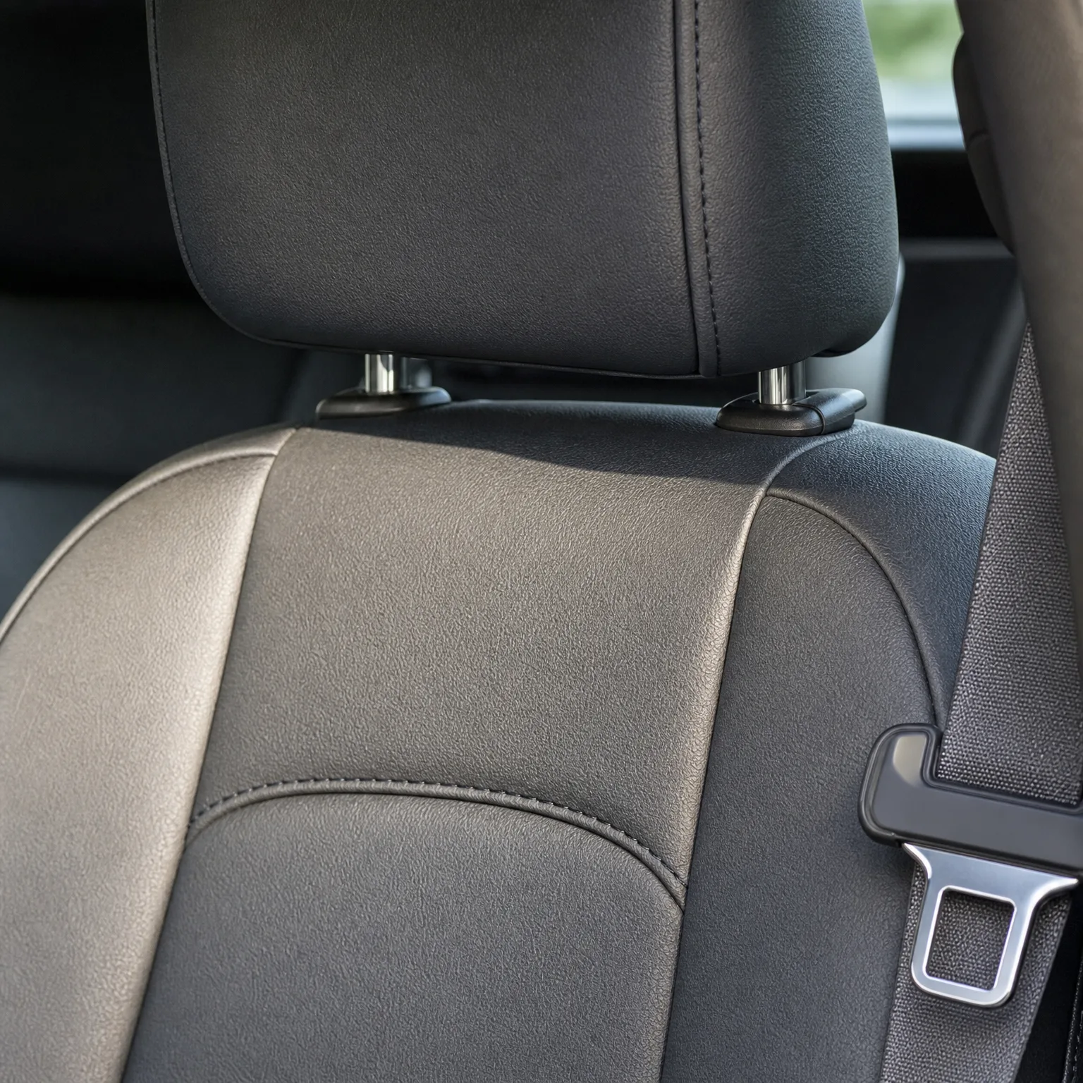 Close-up of a black leather car seat back and headrest with visible stitching, with a seat belt and metal buckle tongue at the right.