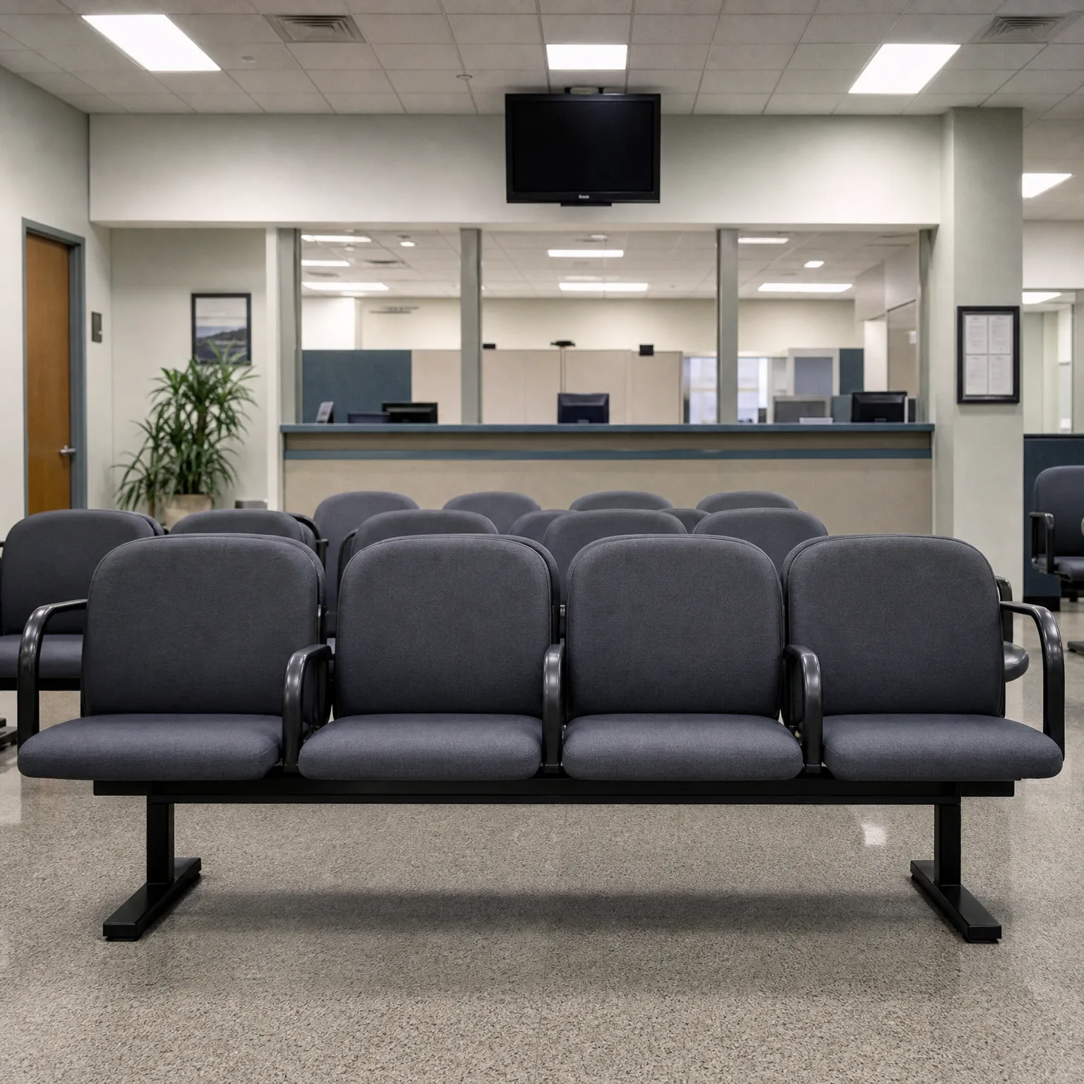 Rows of gray upholstered beam-mounted chairs in an office waiting area with a service counter, wall-mounted flat-screen monitor, potted plant, and speckled floor.